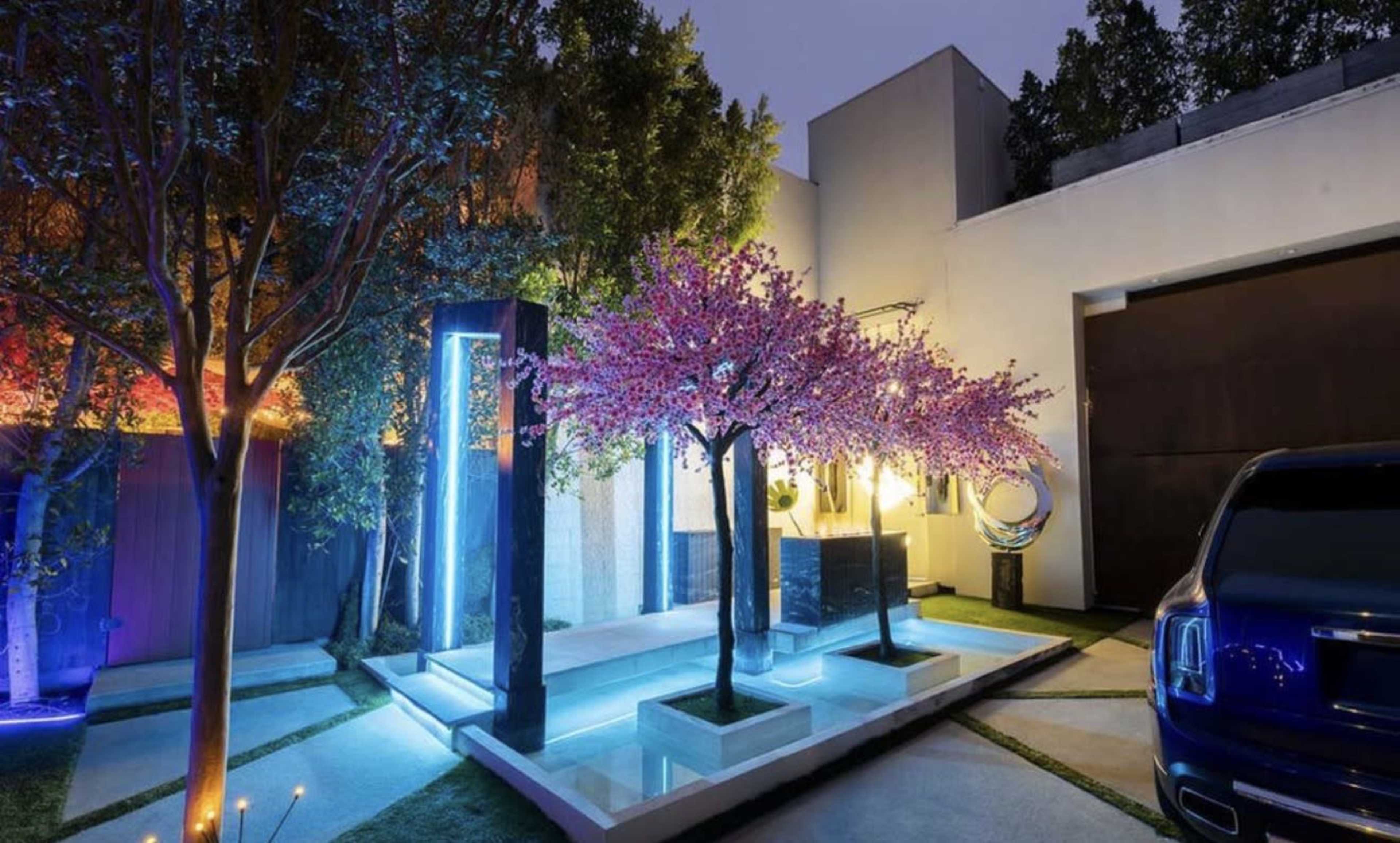 The scene depicts a modern outdoor space featuring a decorative tree with pink blossoms, illuminated by blue lights, alongside a sleek vehicle parked near the entrance of a contemporary building.