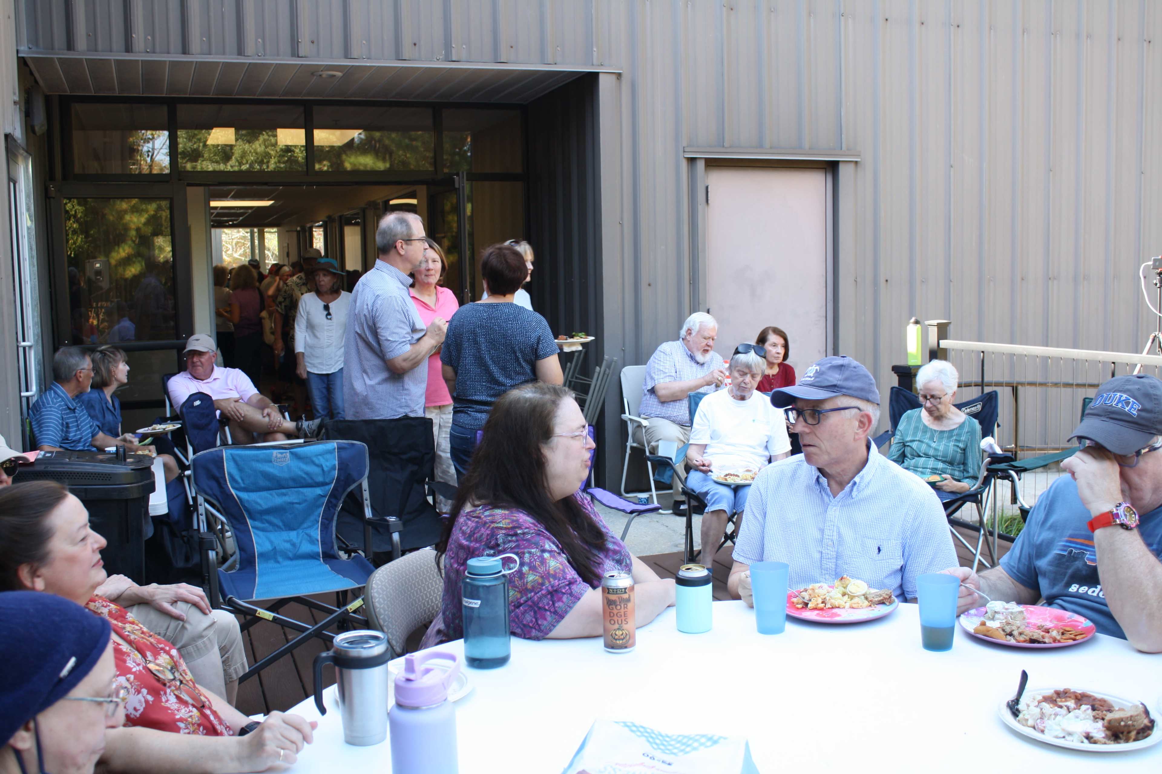 A group of people is gathered outdoors at a social event, sharing food and conversation around tables and chairs.