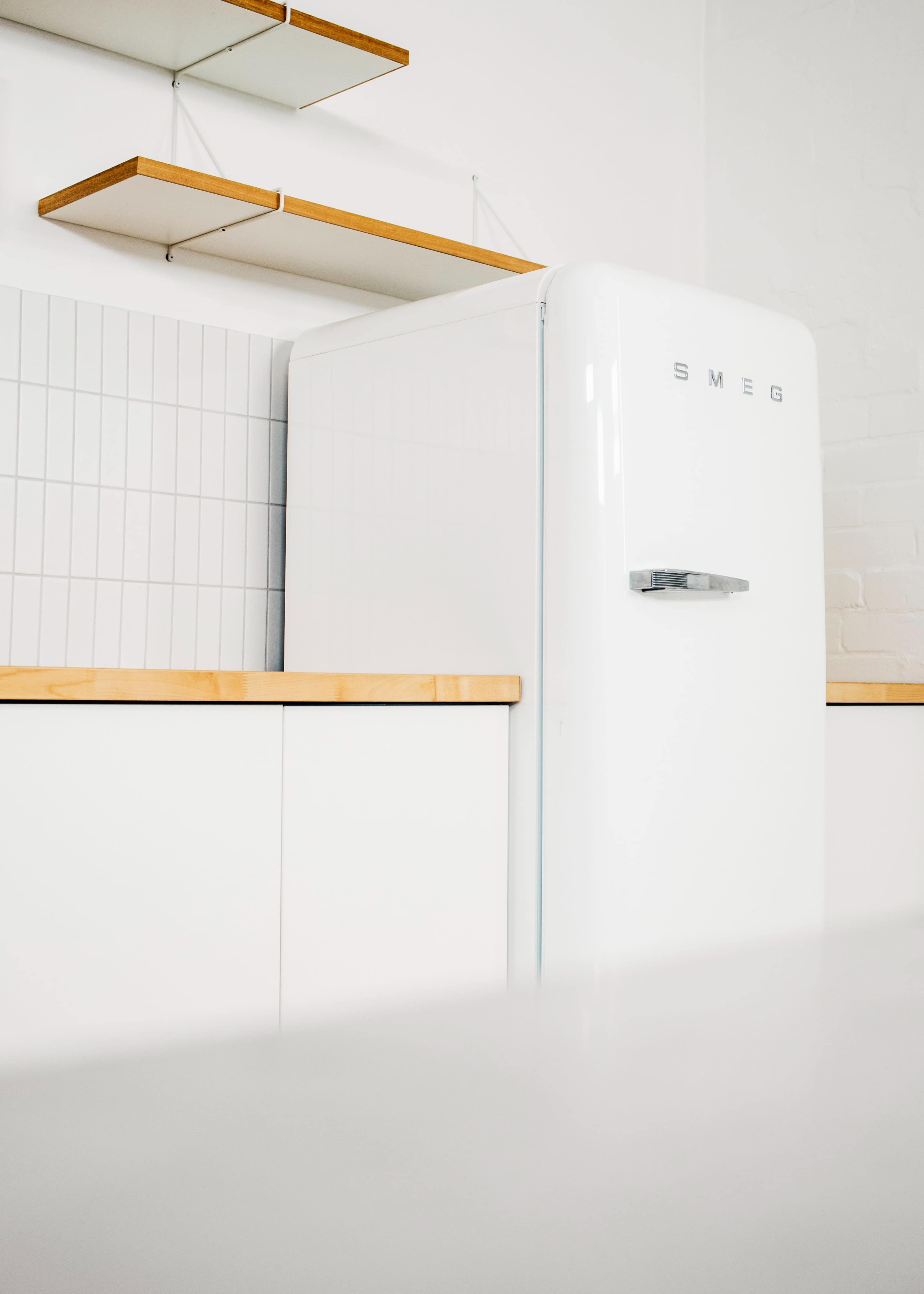 A white SMEG refrigerator next to a minimalist kitchen counter with wooden shelves above.