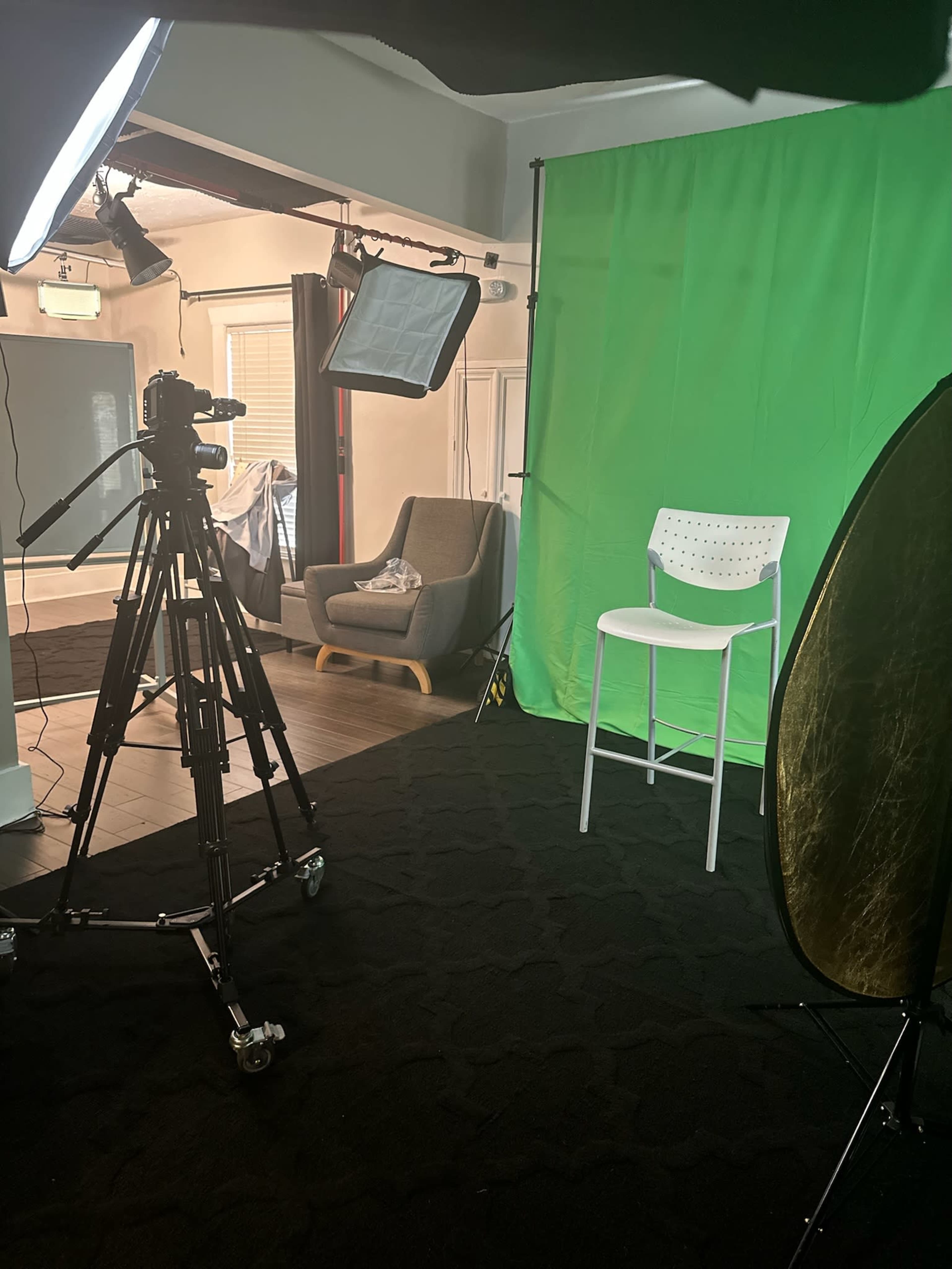 A video recording setup featuring a camera on a tripod aimed at a white chair in front of a green screen, with studio lights positioned to the side.