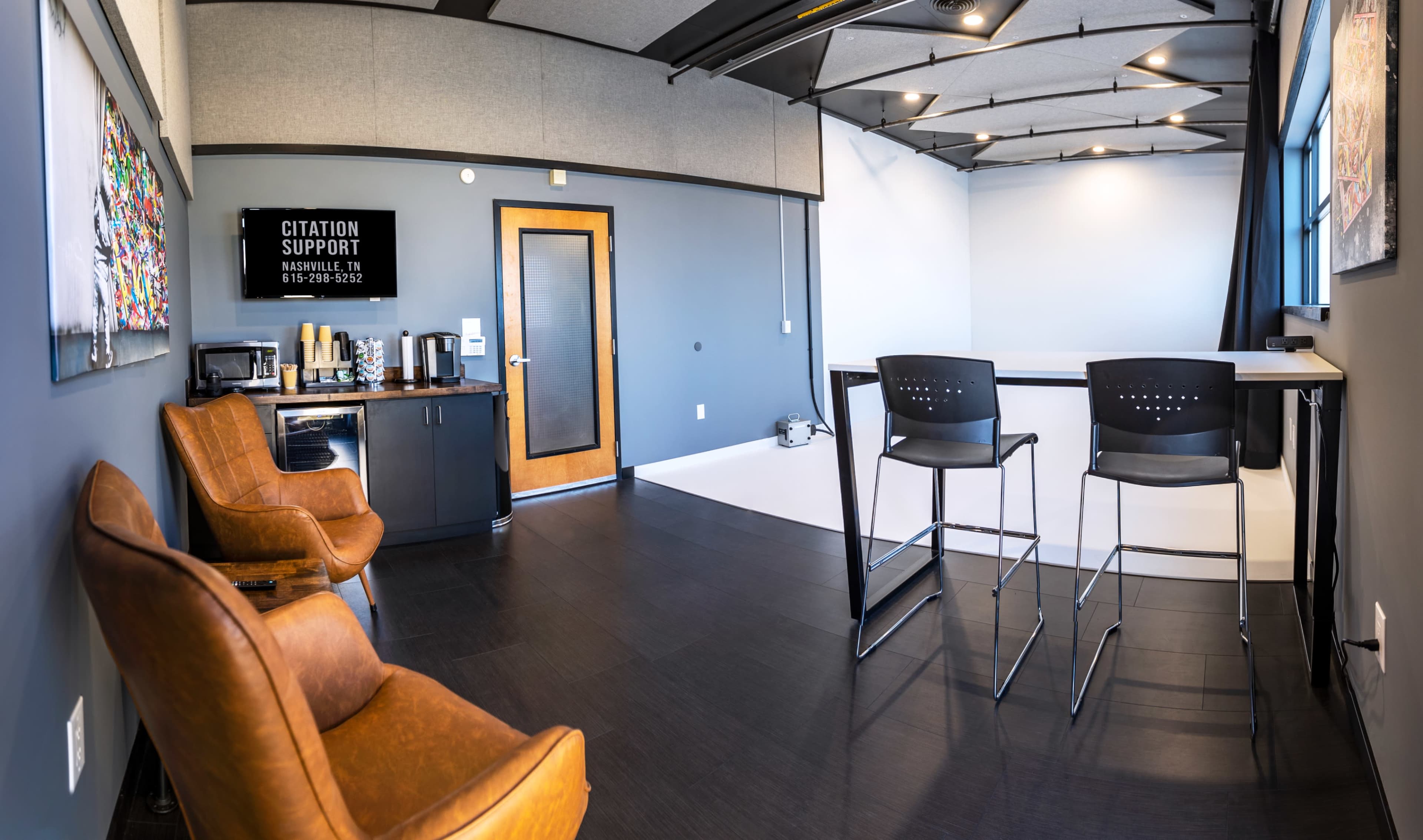 A modern office space featuring a small kitchenette area with a coffee station, two high chairs at a standing table, and a backdrop for photography or video production.