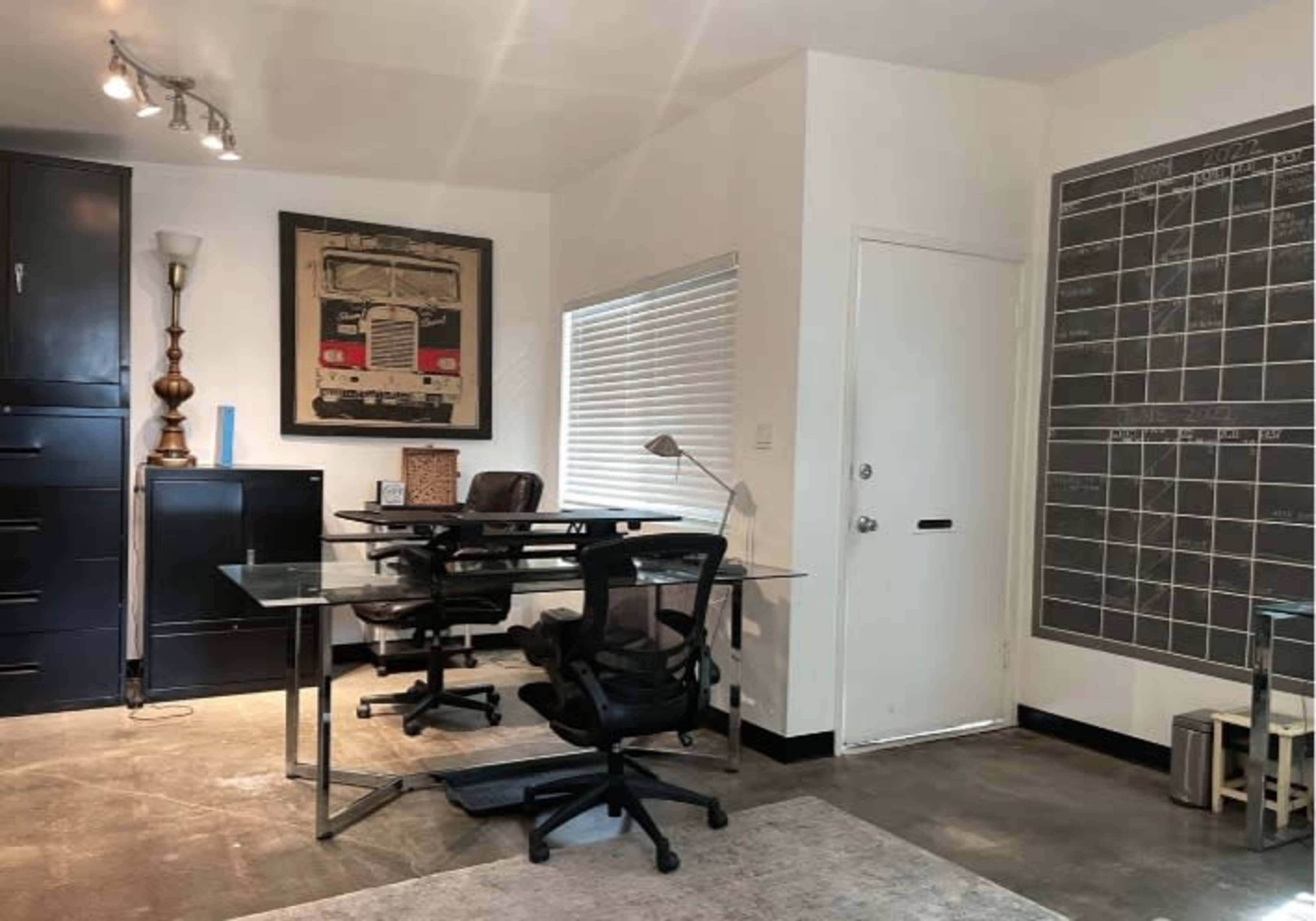 A modern office space with two black office chairs, a glass desk, a large black storage cabinet, and a wall-mounted chalkboard.
