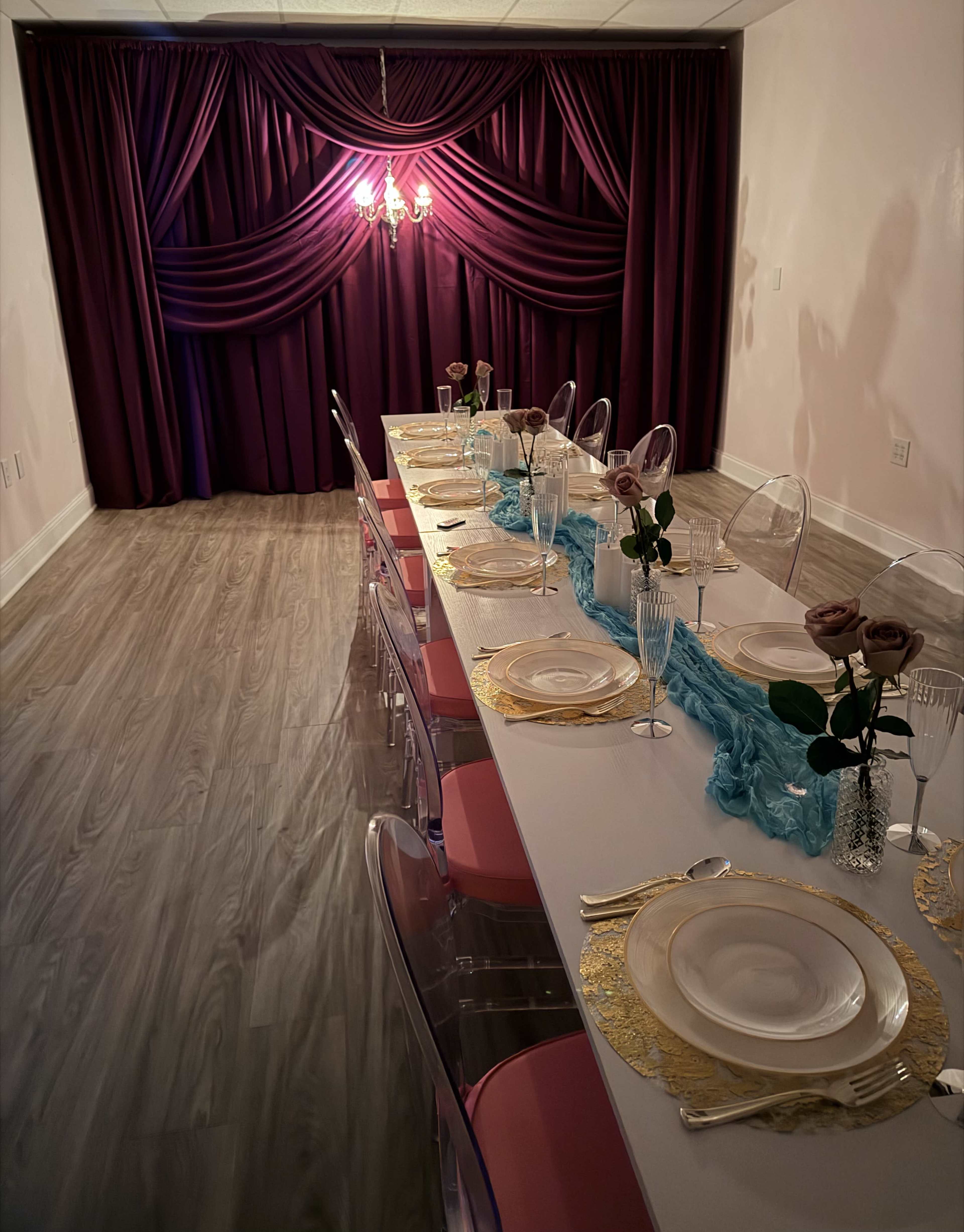 A long dining table is set with elegant tableware and surrounded by clear chairs, faced by a backdrop of draped purple fabric and a chandelier.