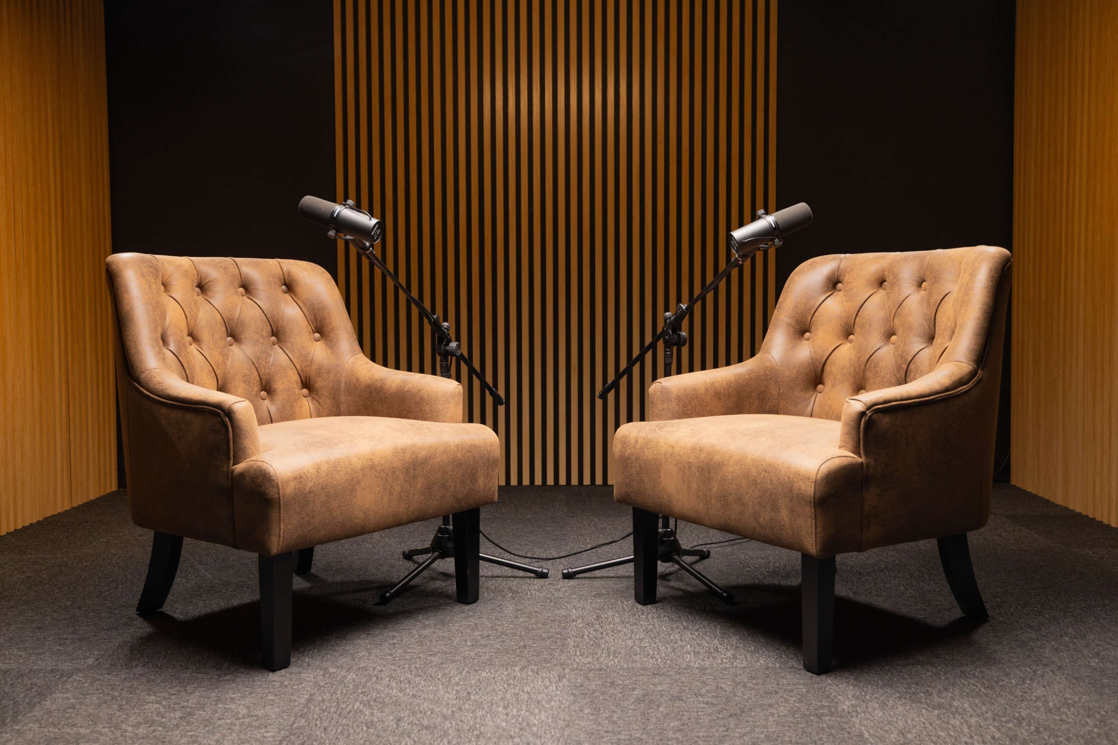 Two tufted brown armchairs are positioned facing each other in a recording studio, each equipped with a microphone on a stand.