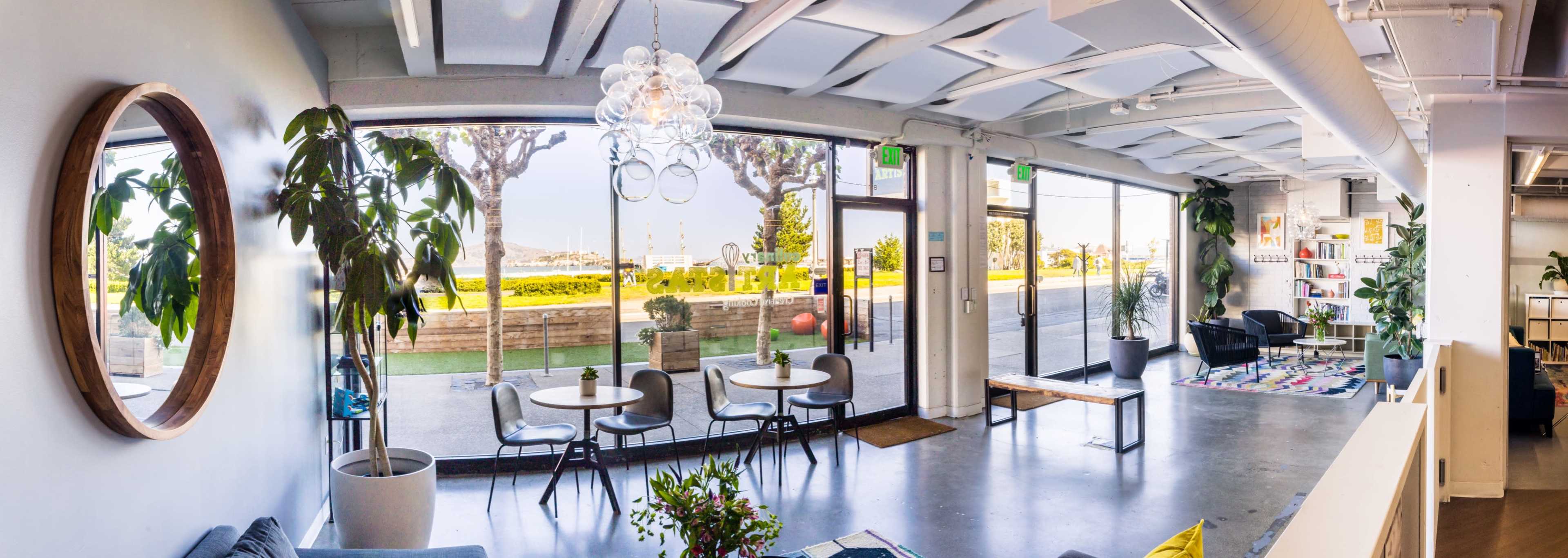 A panoramic view of a modern interior space featuring a circular mirror, indoor plants, tables and chairs, large windows overlooking a landscaped area, and a glimpse of the outdoors.