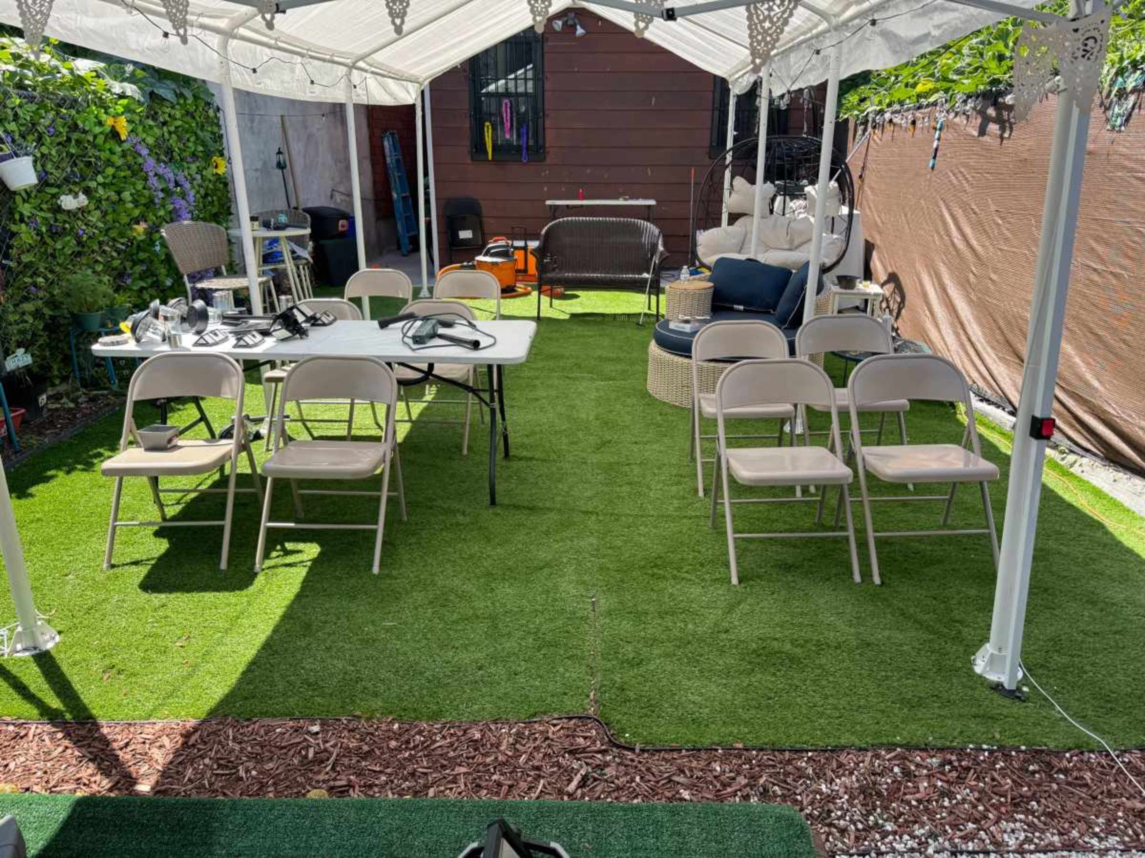 A covered outdoor area features a large table with several folding chairs arranged around it, set on artificial grass, with seating and decorations in the background.
