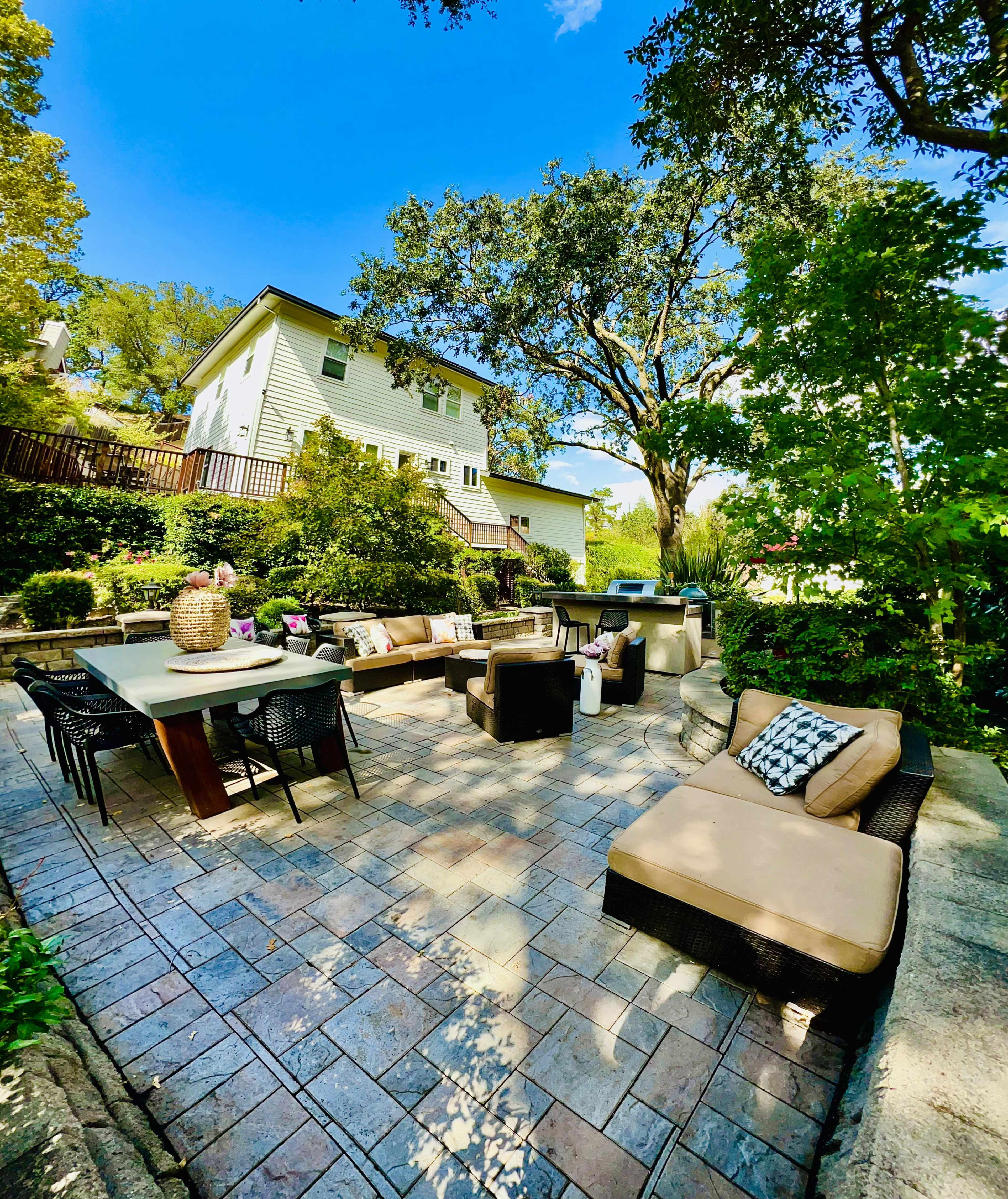 The image shows a patio area featuring a dining table, several lounge chairs, and a house in the background, surrounded by greenery.