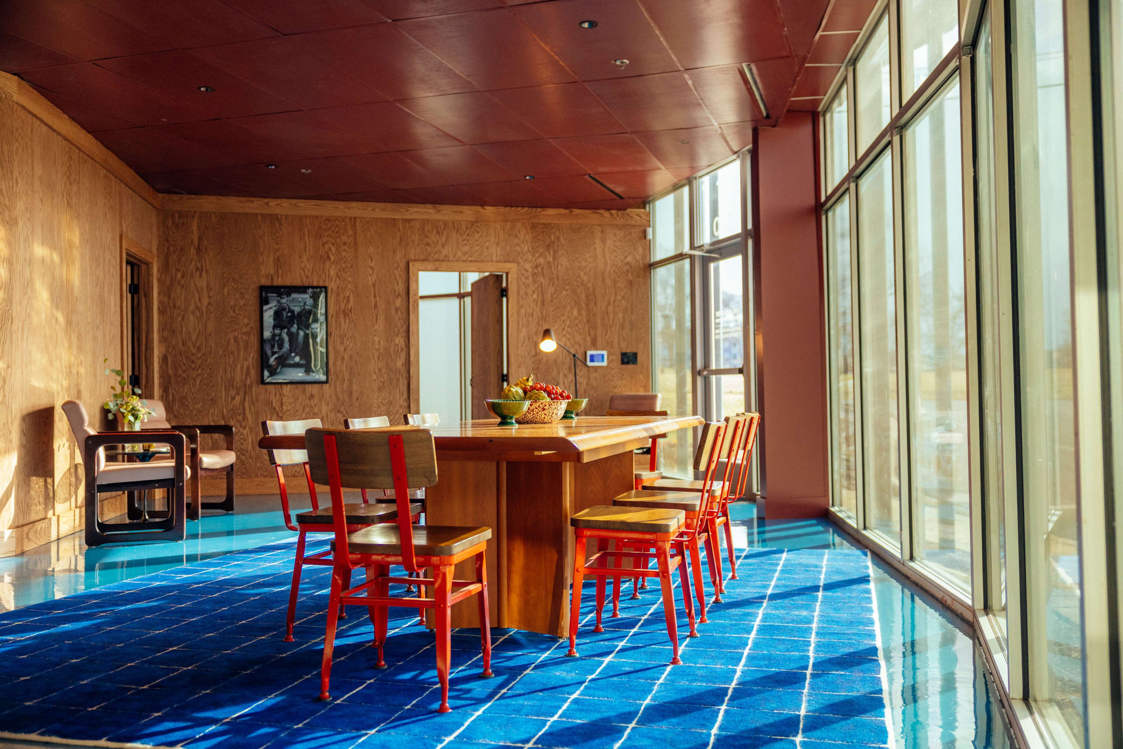 The image shows a modern dining area with a wooden table surrounded by red chairs, set against a backdrop of large windows allowing natural light to flood the space.