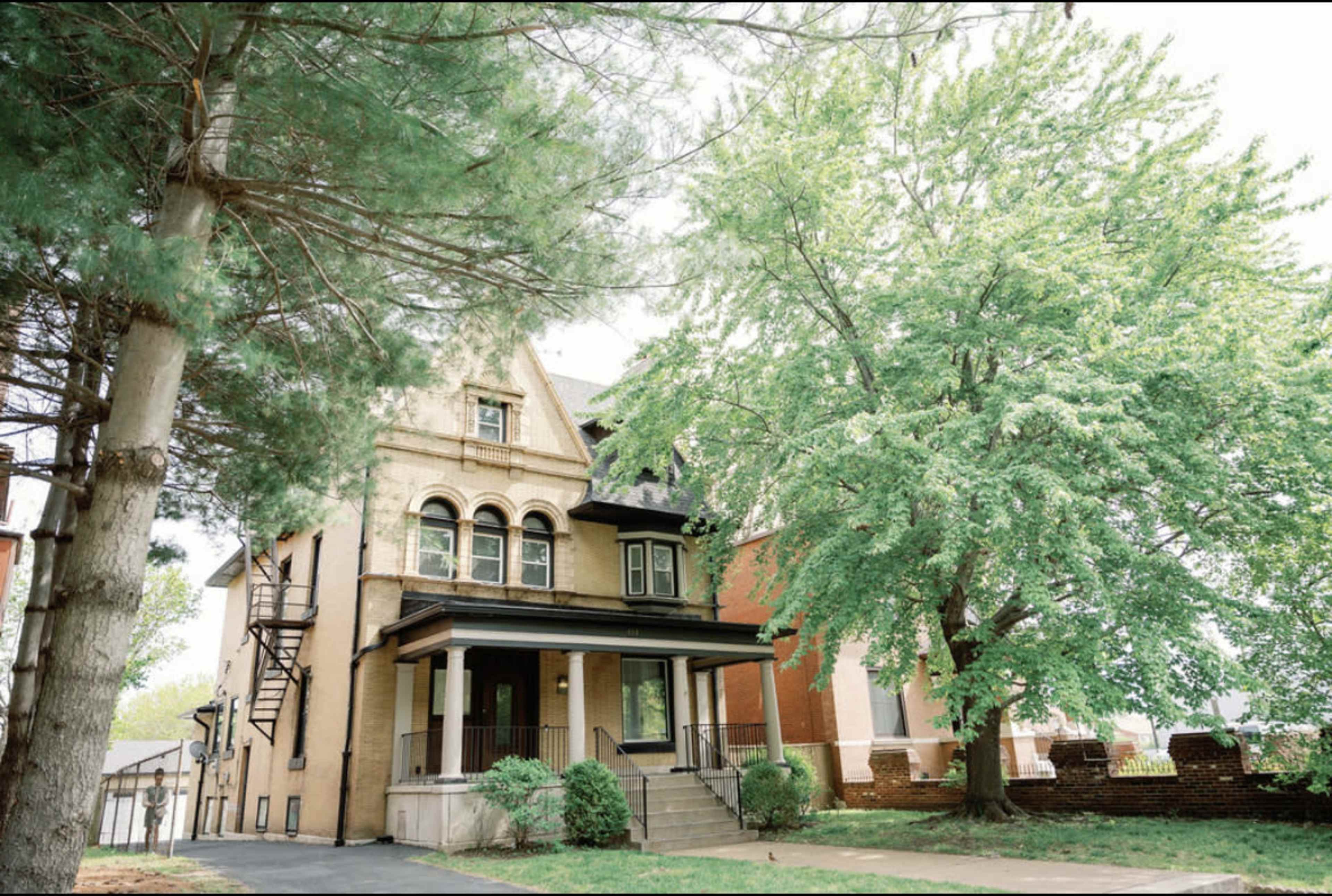 A large, multi-story house with a distinctive architectural style is situated next to a leafy tree and surrounded by well-maintained landscaping.
