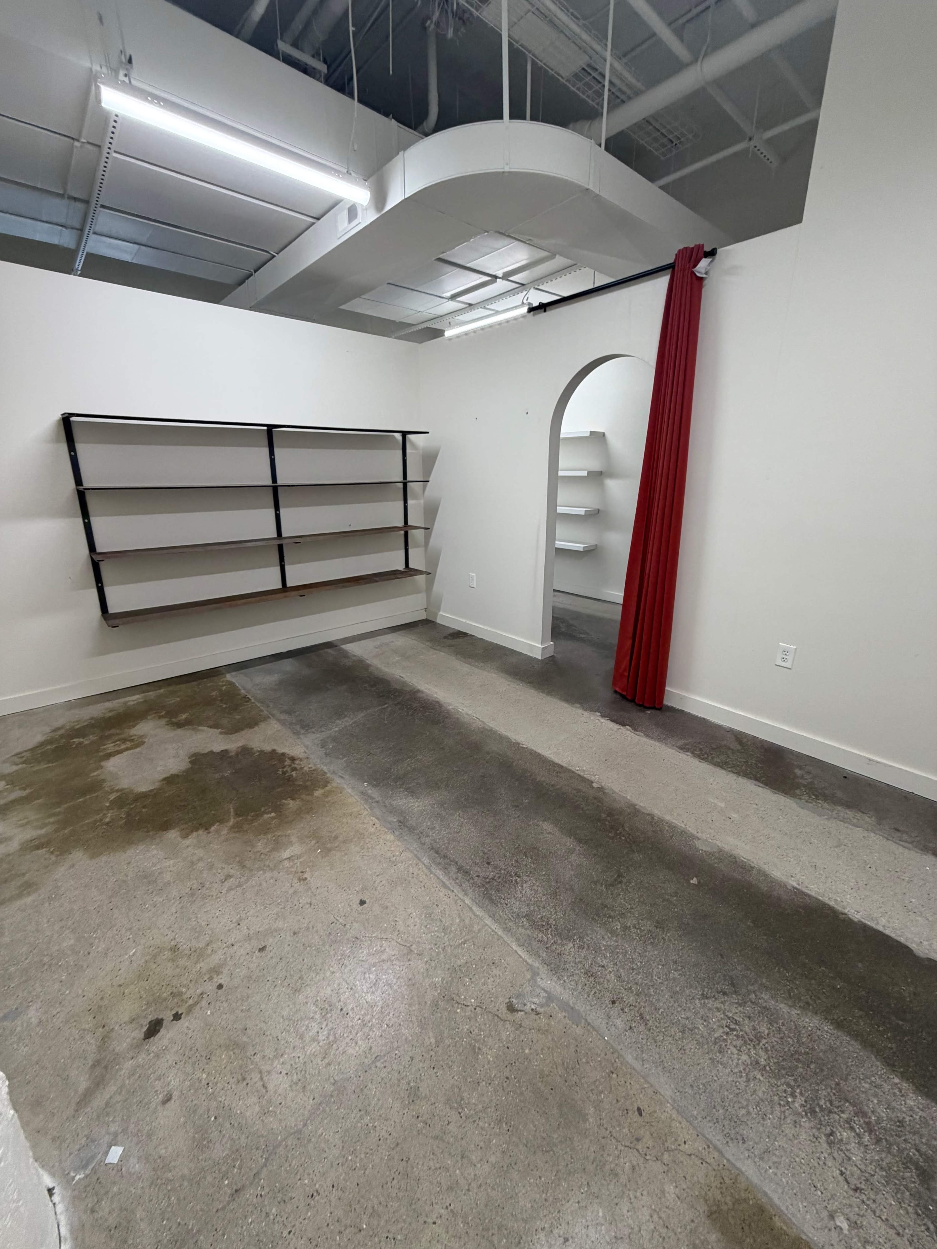 An empty room features gray concrete flooring, white walls, and a curved archway, with shelves mounted on one wall and a red curtain hanging nearby.