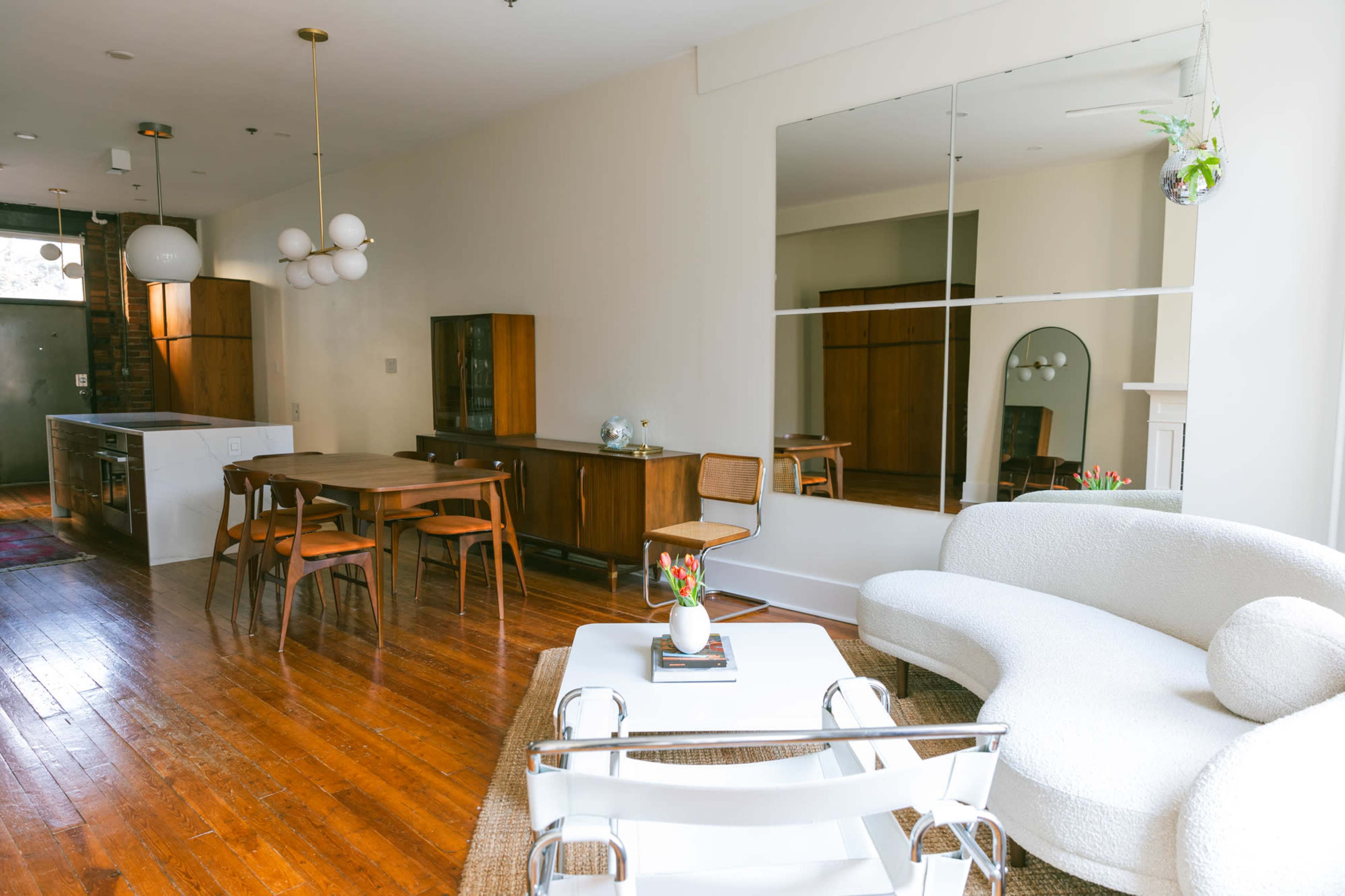 A modern living space with wooden floors, a small dining area, and a large mirror reflecting a bright, open layout.