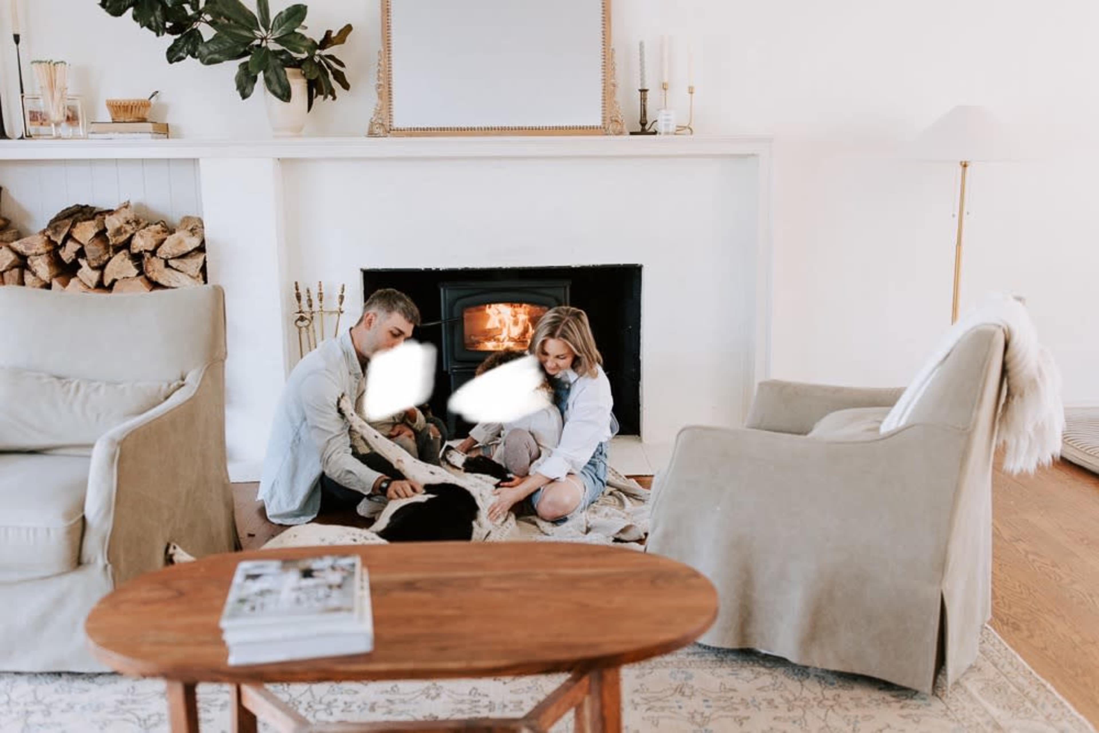 A man and a woman sit on the floor near a fireplace, playing with a dog while surrounded by blankets in a cozy living room.