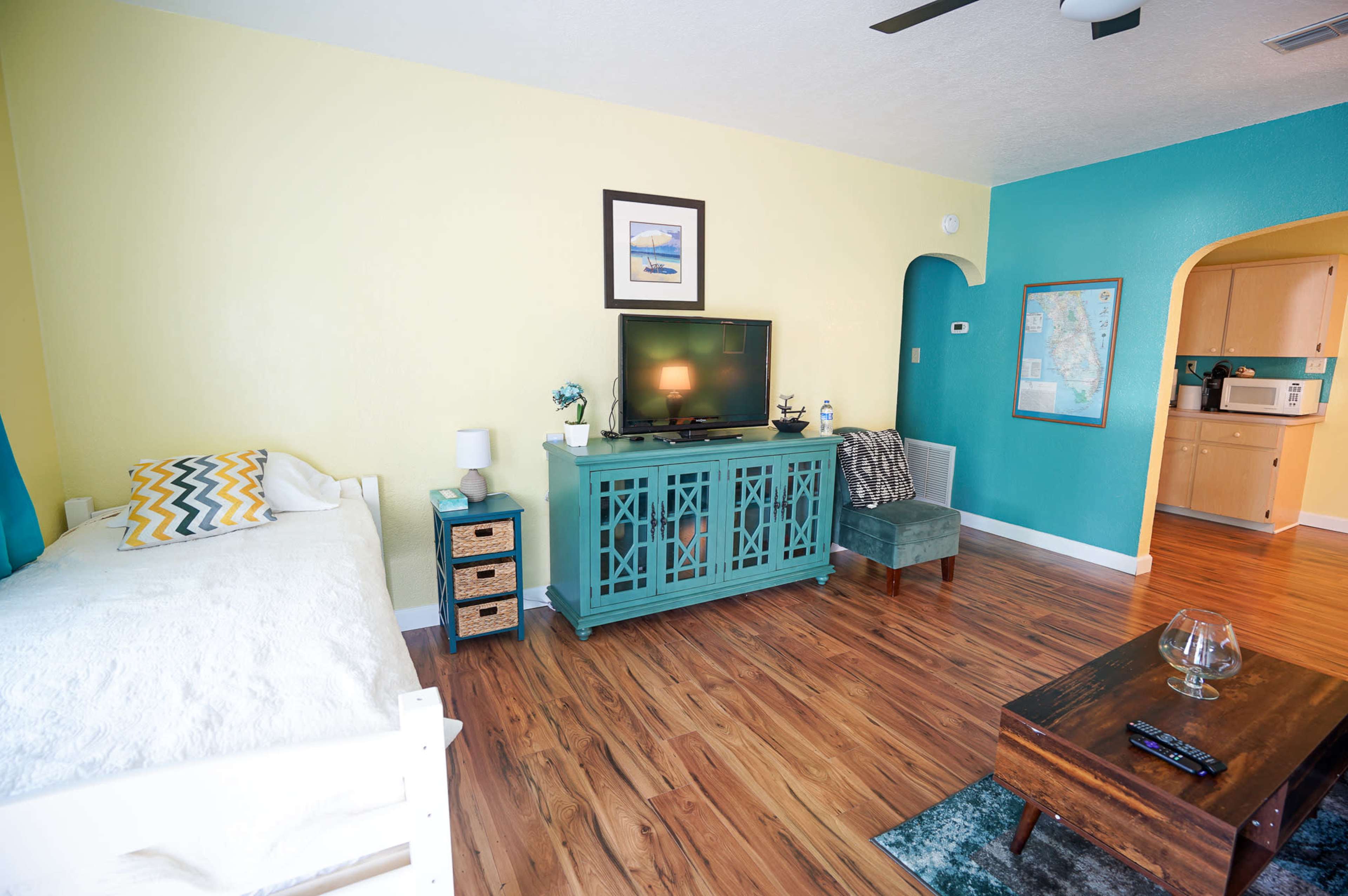 A brightly colored living room featuring a television on a teal cabinet, a white bed, and a small dining area with an archway leading to a kitchen.