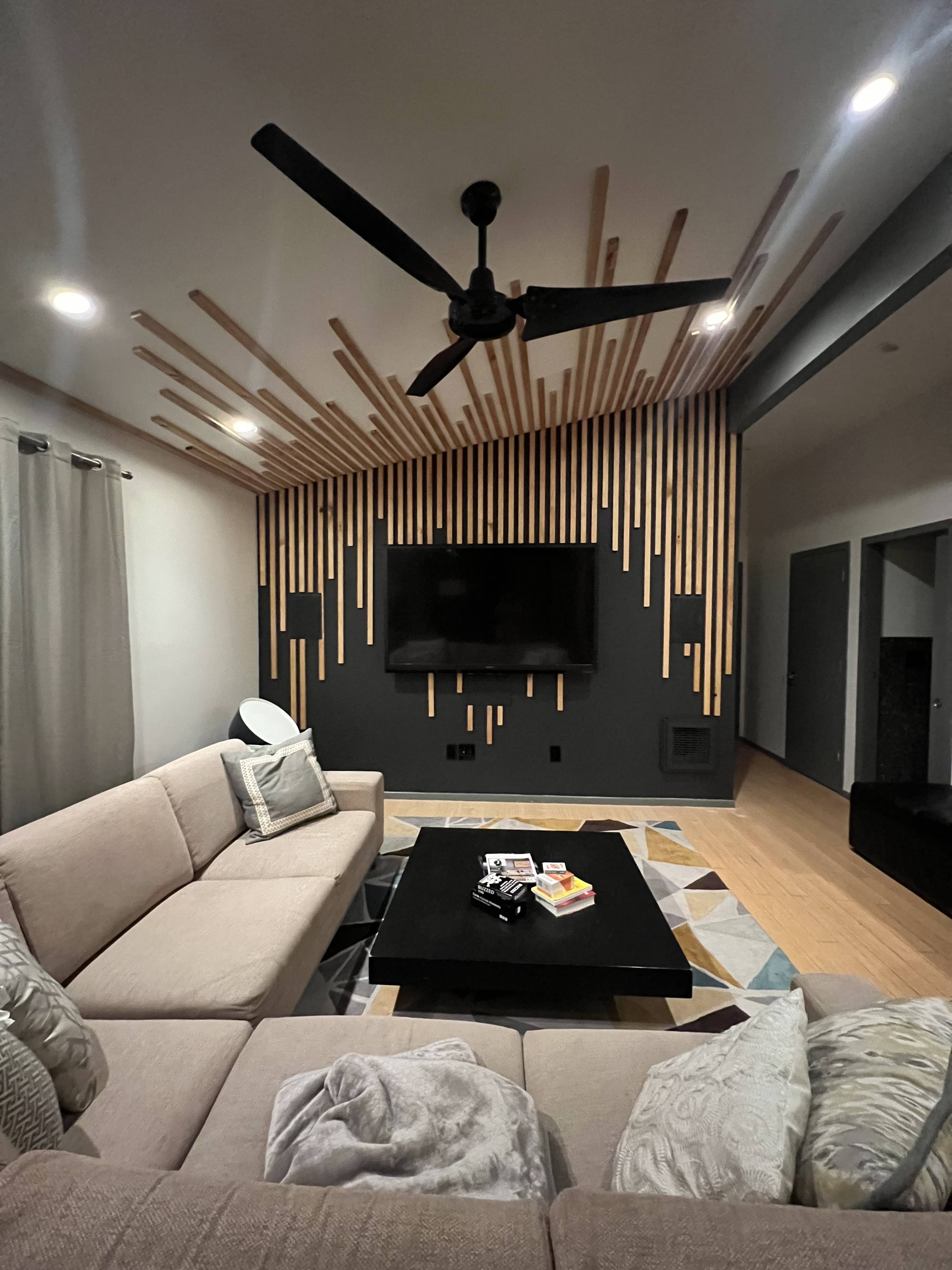 A modern living room with a light-colored sectional sofa, a black coffee table, and a wall decorated with vertical wooden slats behind a mounted television.