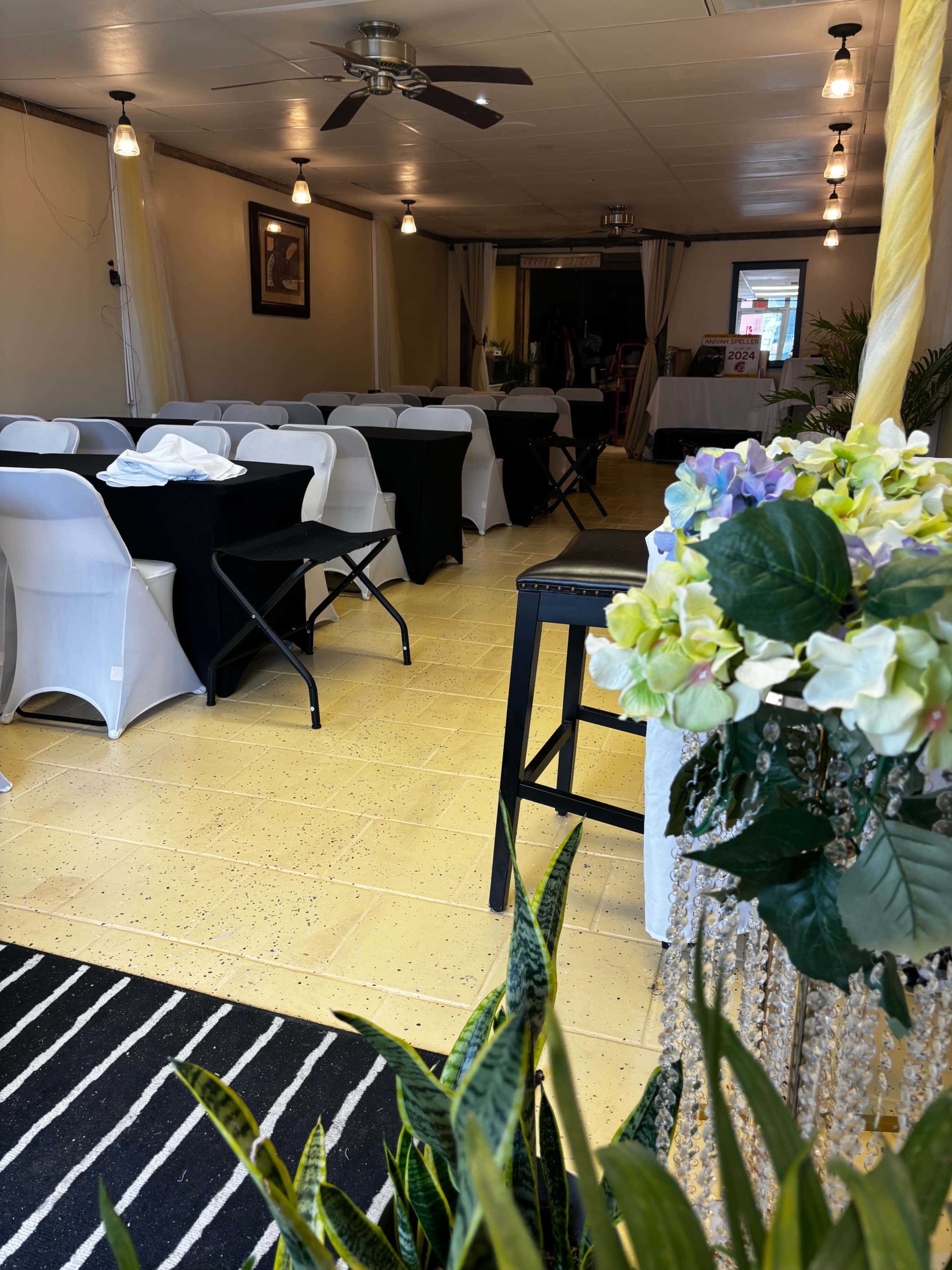 The image shows a partially decorated indoor event space with tables covered in black and white linens, a striped rug, and plants arranged in the foreground.