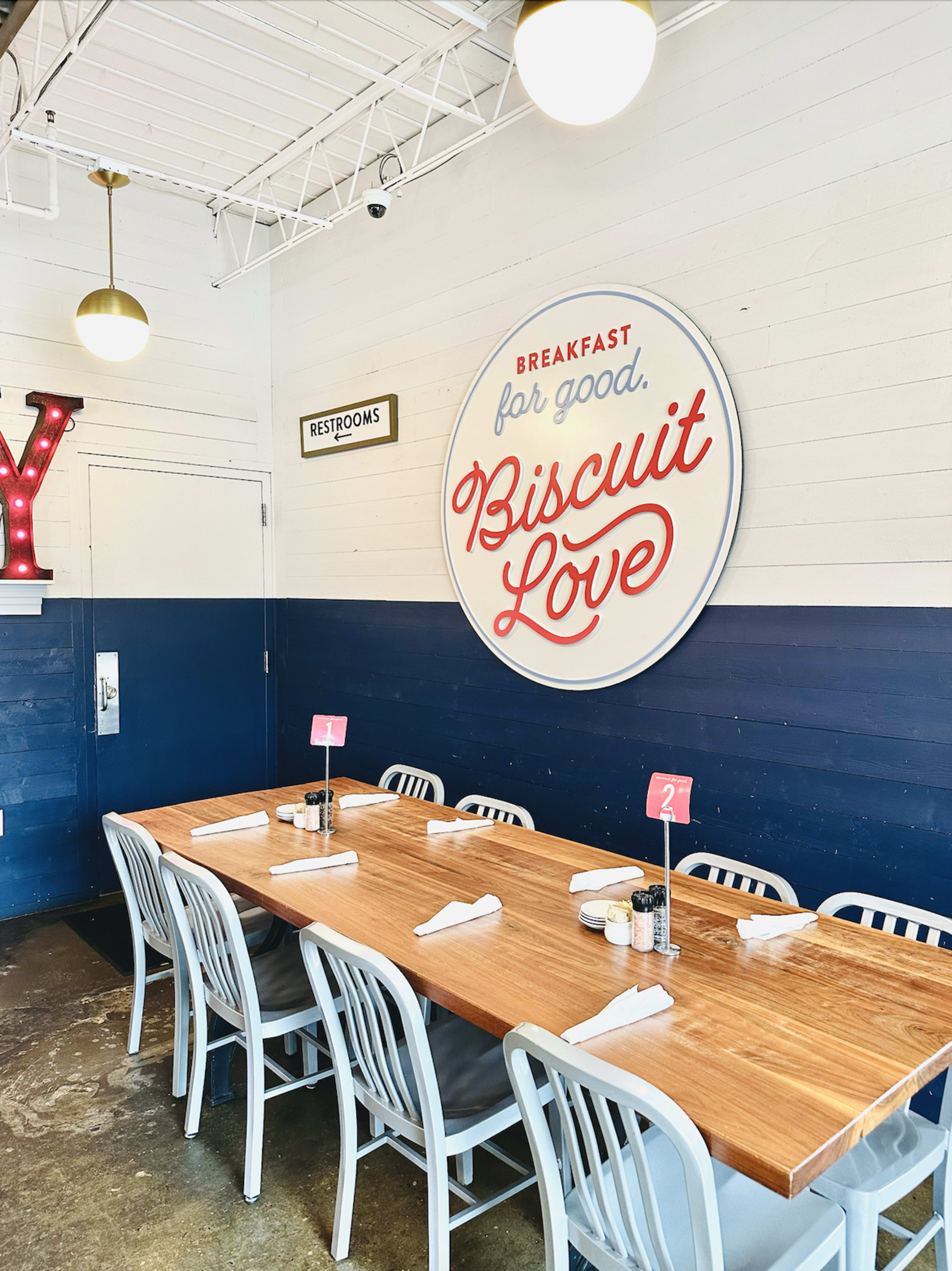 A wooden table with white chairs is set in a casual dining area featuring a wall sign that reads "Biscuit Love" and indicates breakfast offerings.