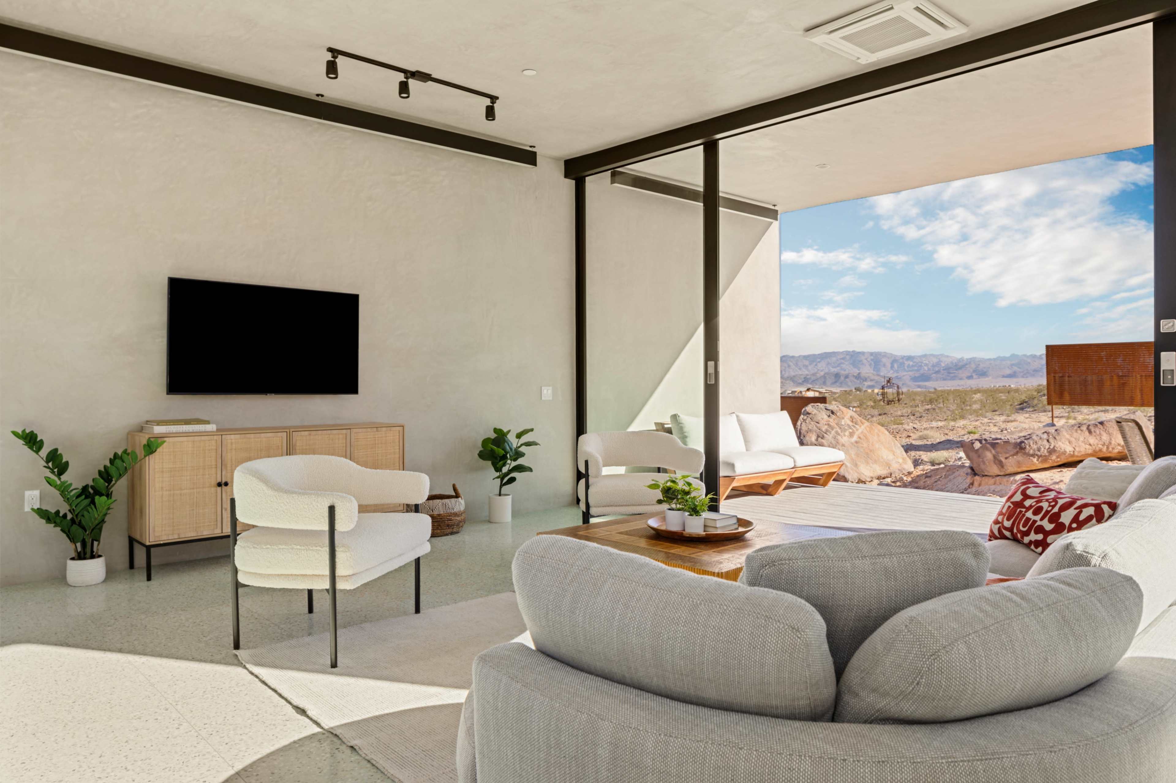 A modern living room features a comfortable seating area, a TV on a cabinet, and large windows offering a view of a rocky landscape under a partly cloudy sky.