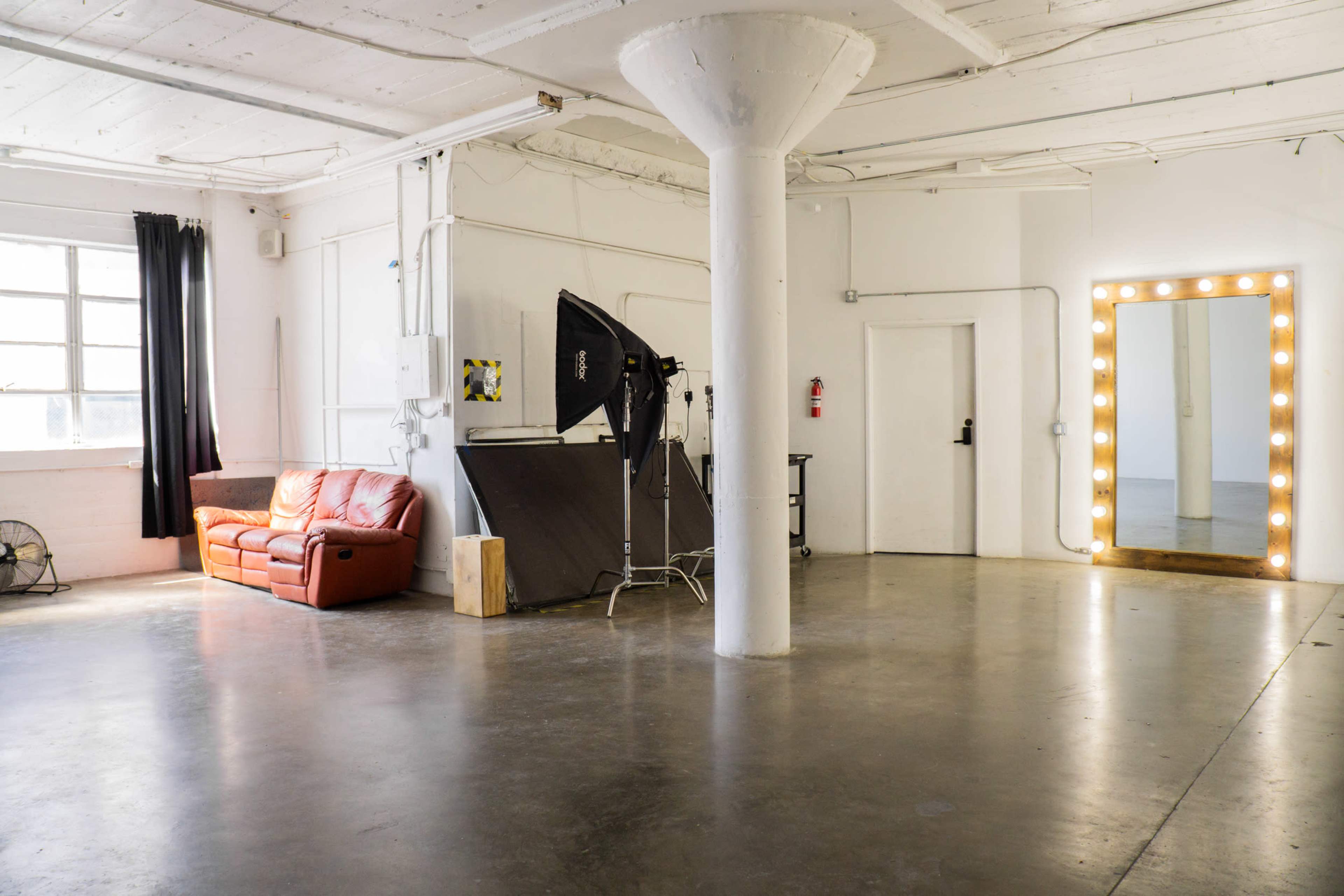 The image shows a large, empty room featuring a red leather couch, a standing mirror with lights, and photography equipment set up in one corner.