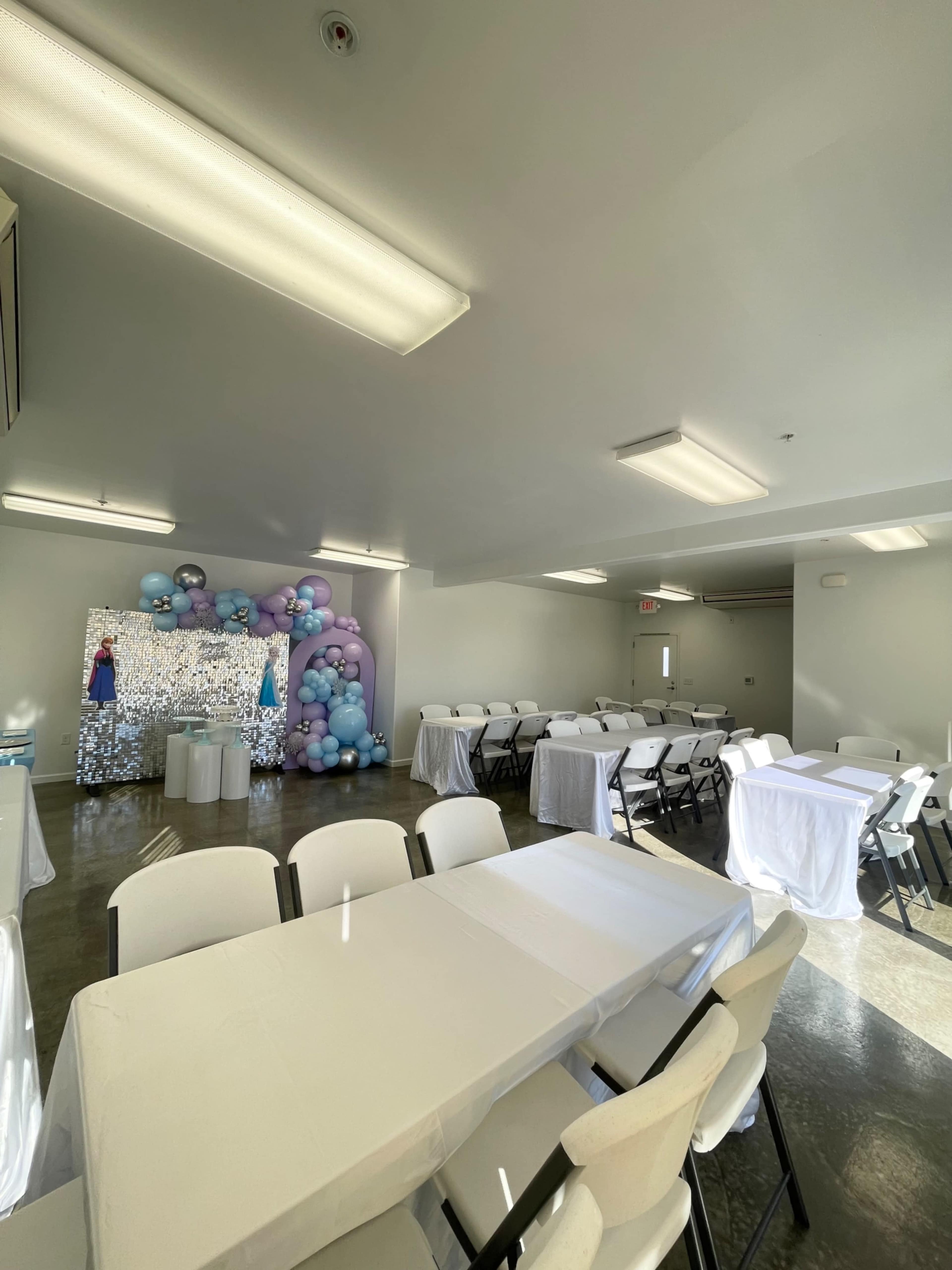 A spacious event room with white tables and chairs set up, featuring a large balloon arch and a decorative backdrop at one end.
