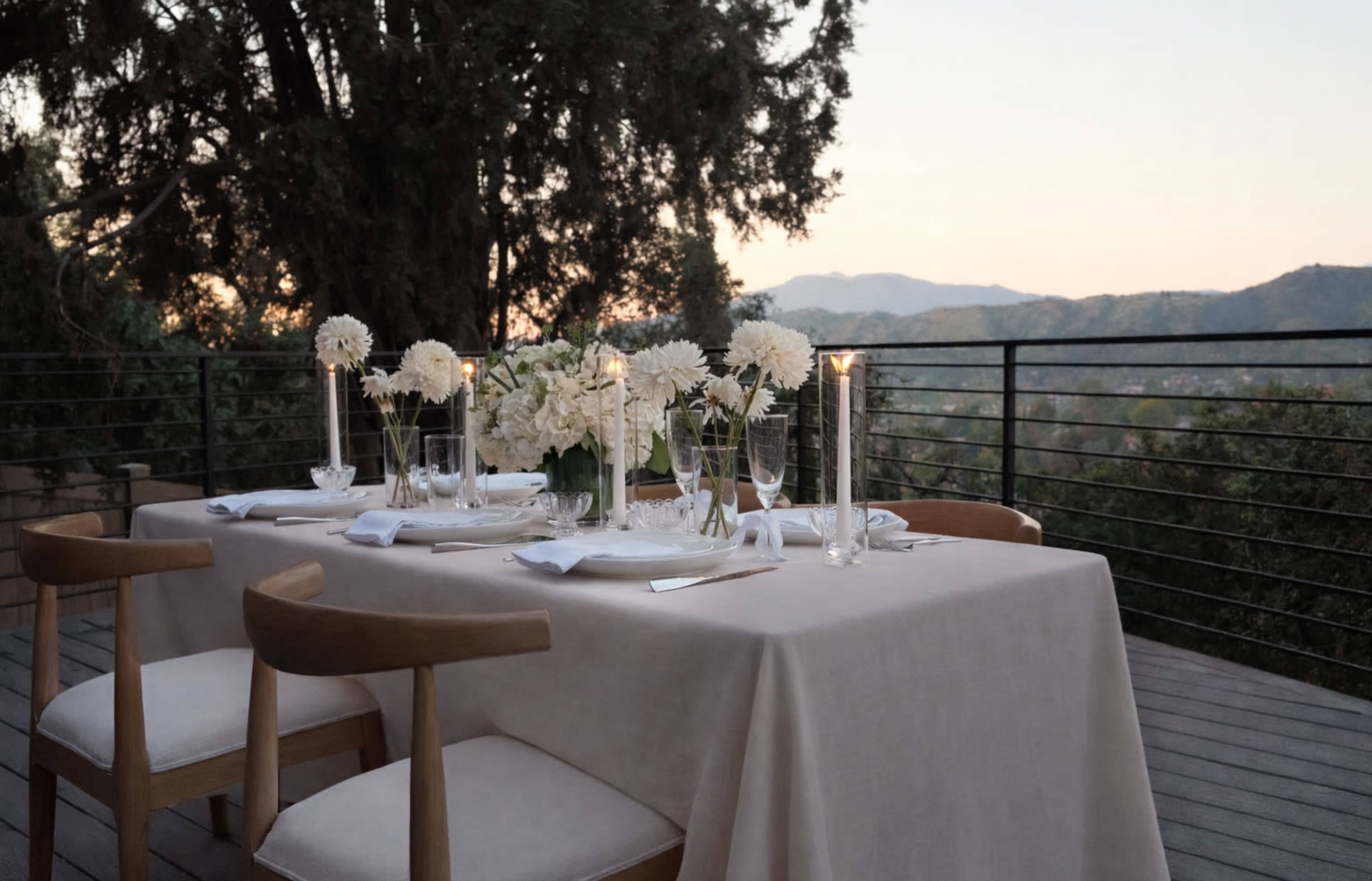 A dining table is set outside with white flowers and candles, overlooking a mountainous landscape at sunset.