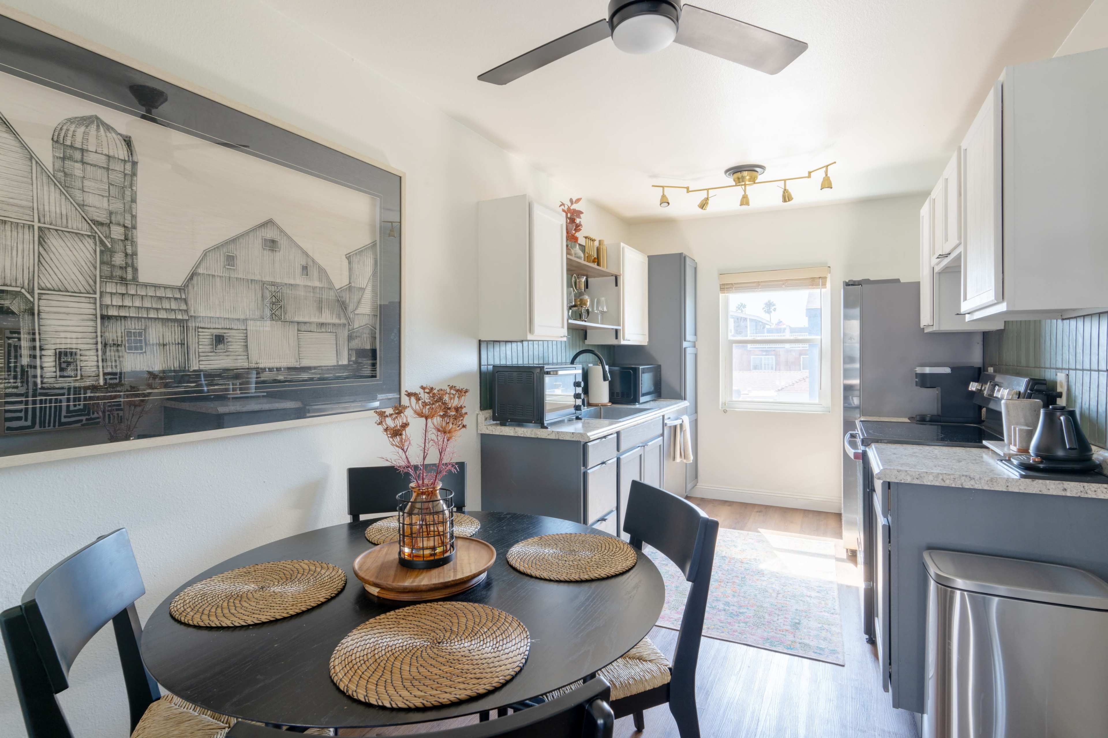 A modern kitchen features a round table with place settings, a black ceiling fan, and an artistic farmhouse print on the wall.
