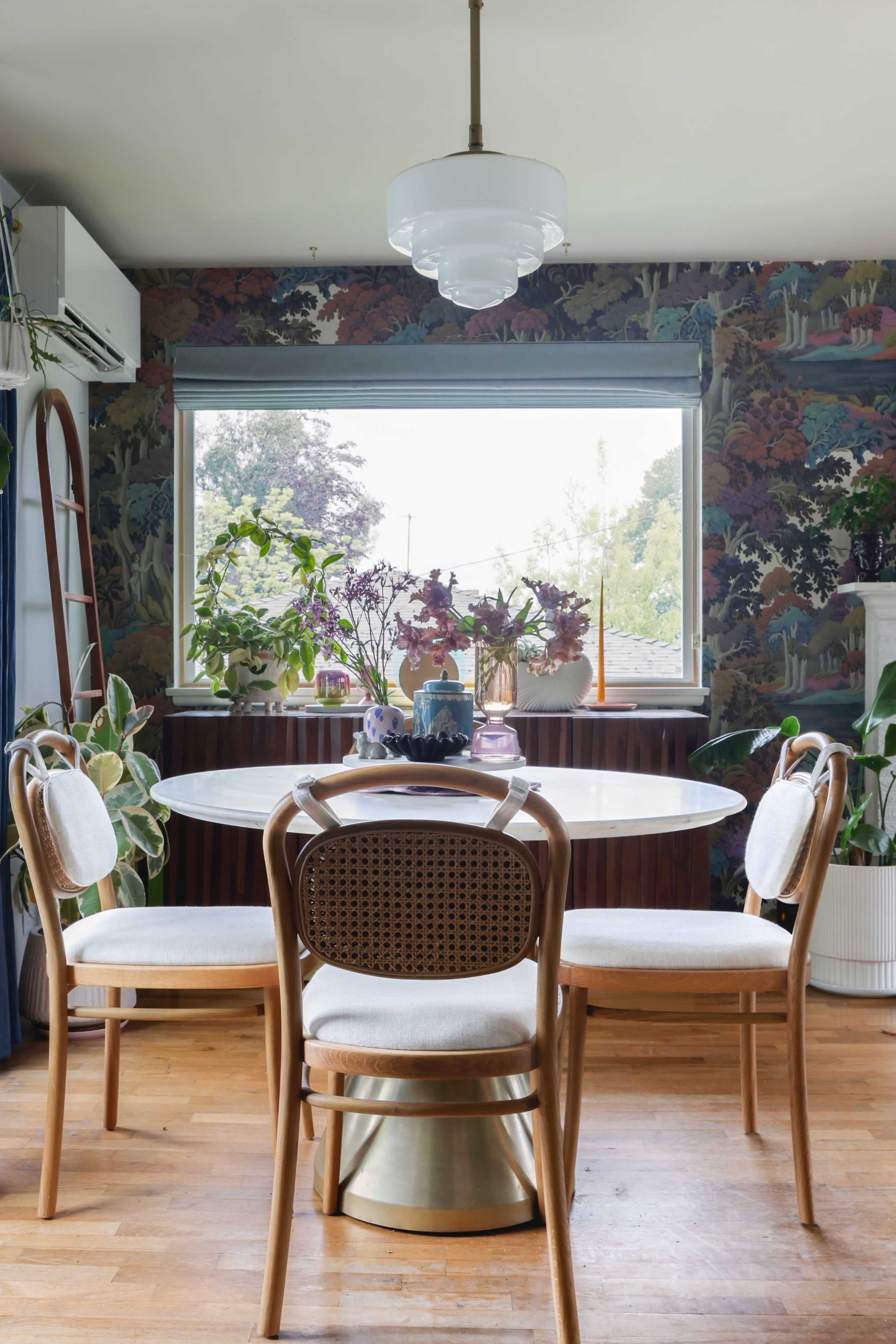 A round dining table is surrounded by four chairs in a brightly lit room decorated with patterned wallpaper and various indoor plants.