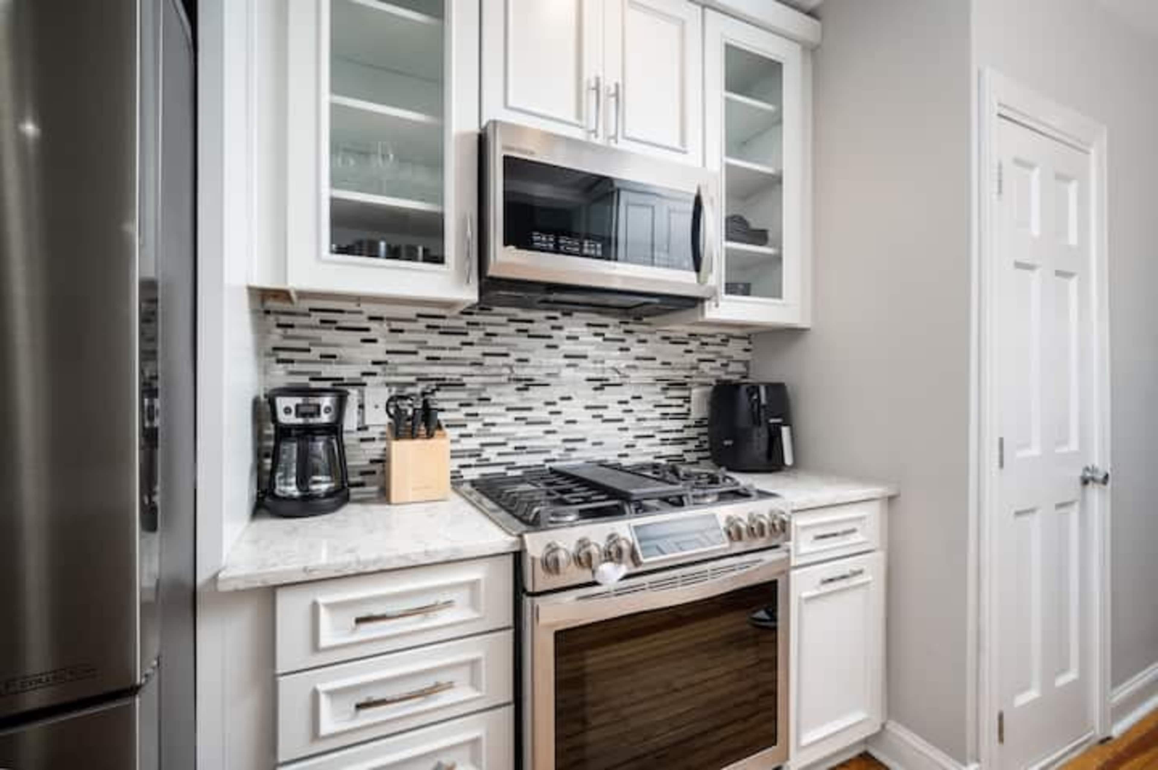 The image shows a modern kitchen with white cabinetry, a stainless steel microwave, and a gas stove beneath a tiled backsplash.