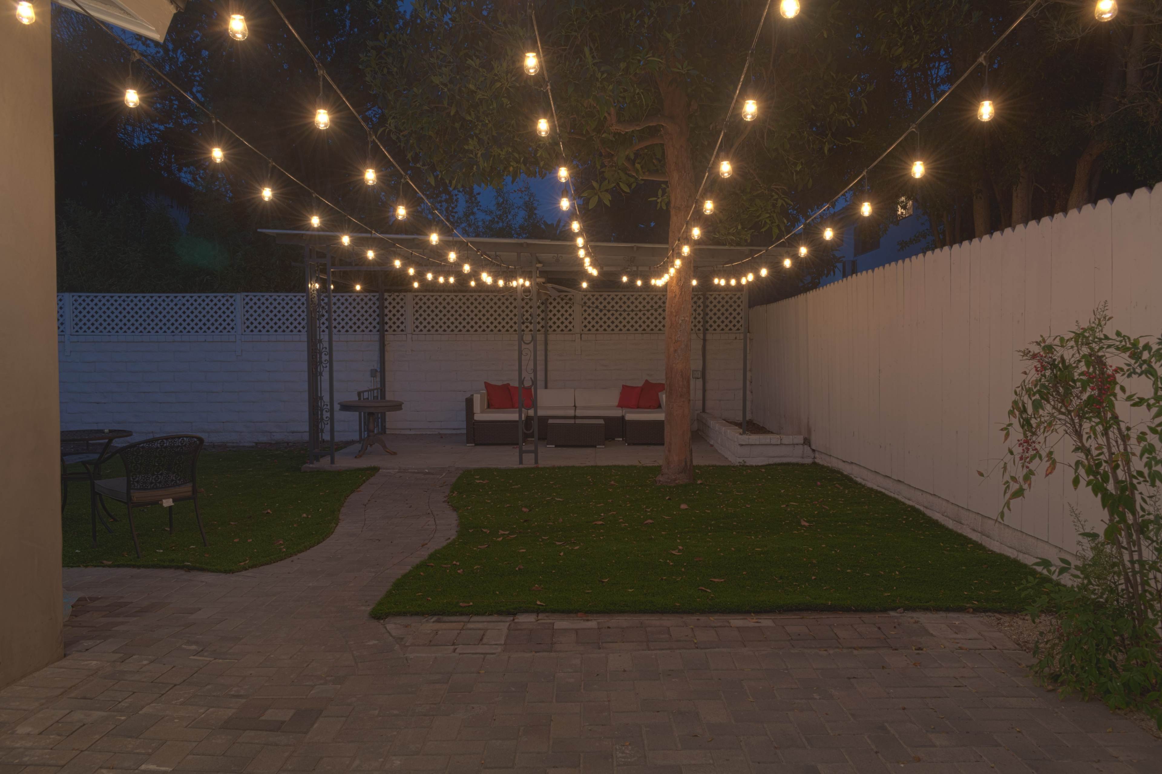 A backyard scene at night, featuring a patio area with string lights overhead, a seating arrangement with red cushions, and a pathway leading to a grassy lawn.
