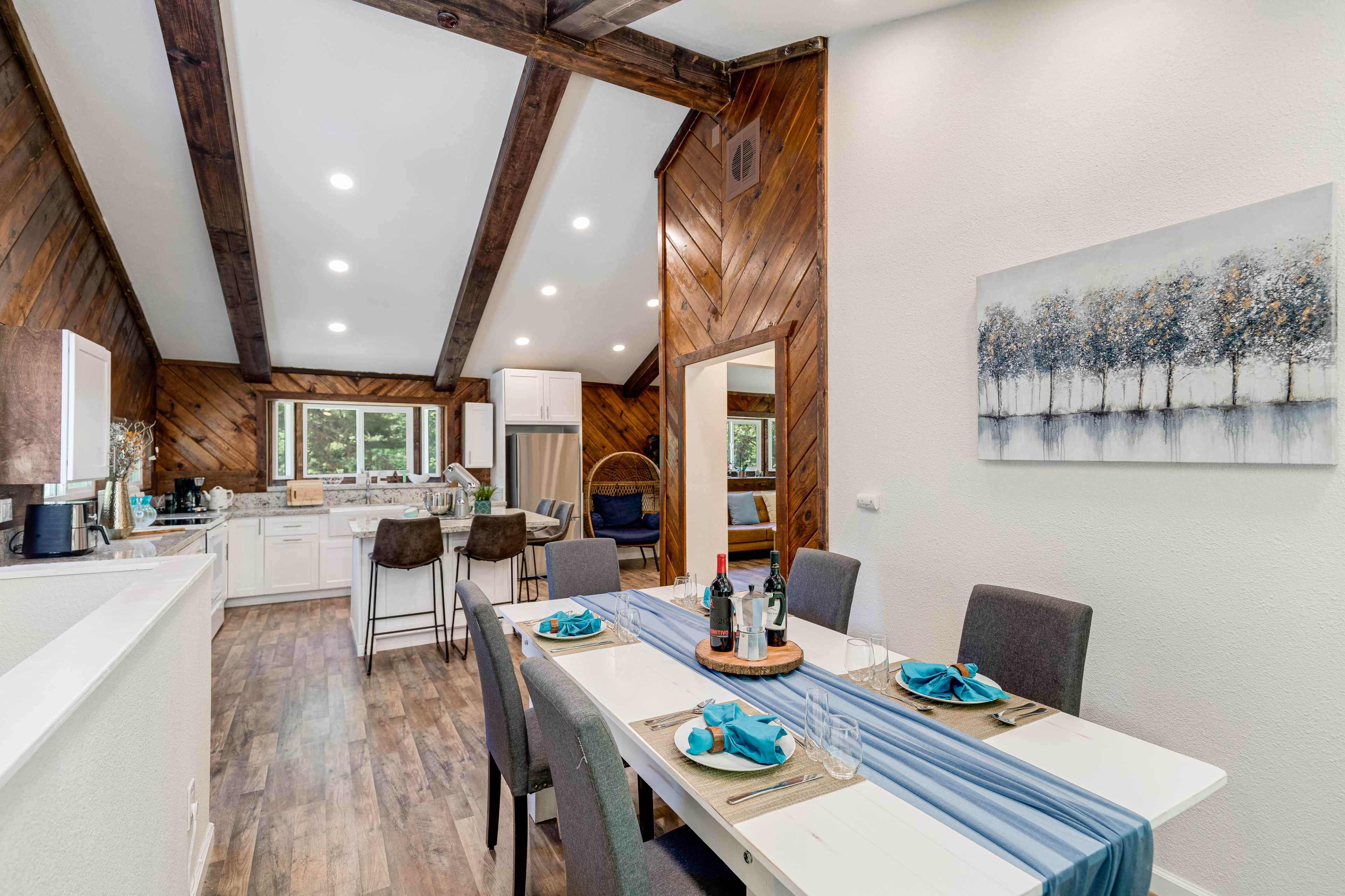 A modern dining area with a table set for a meal, featuring a blue and white table runner, surrounded by gray chairs, and leading into a kitchen with wooden beams and white cabinetry.