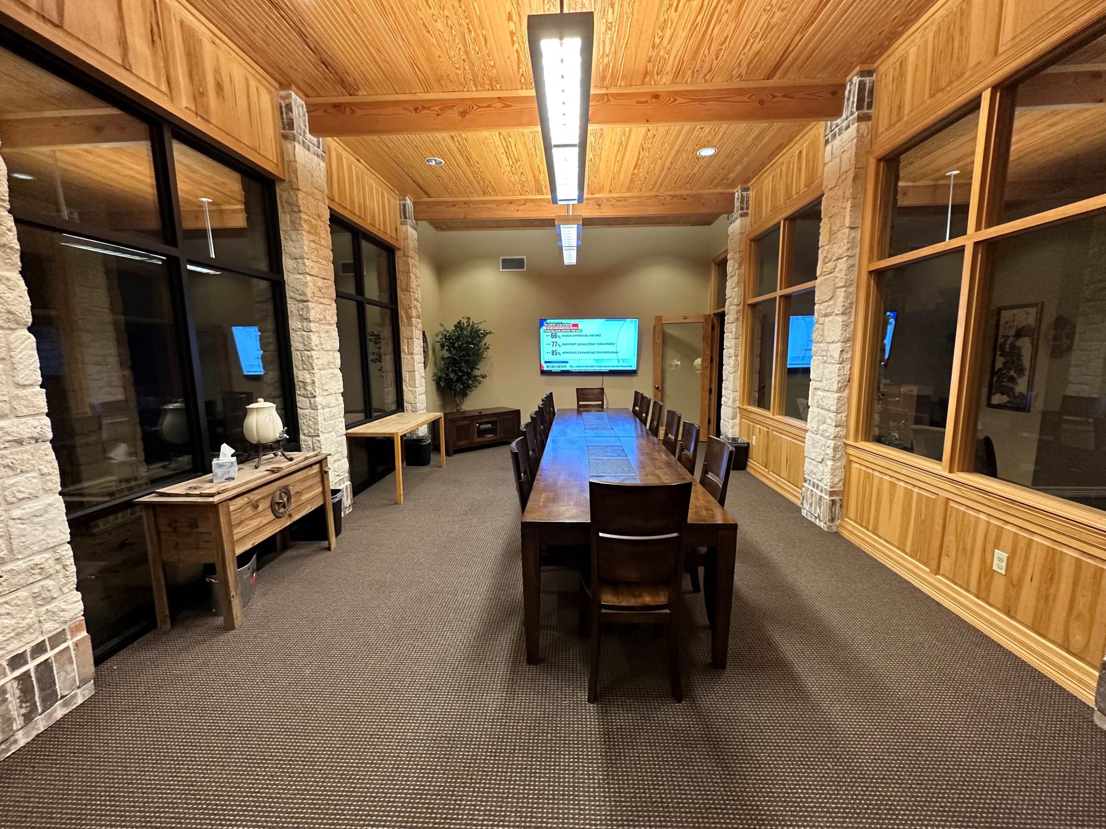 A long wooden conference table with chairs arranged around it in a room featuring large windows and stone walls, illuminated by overhead lights.