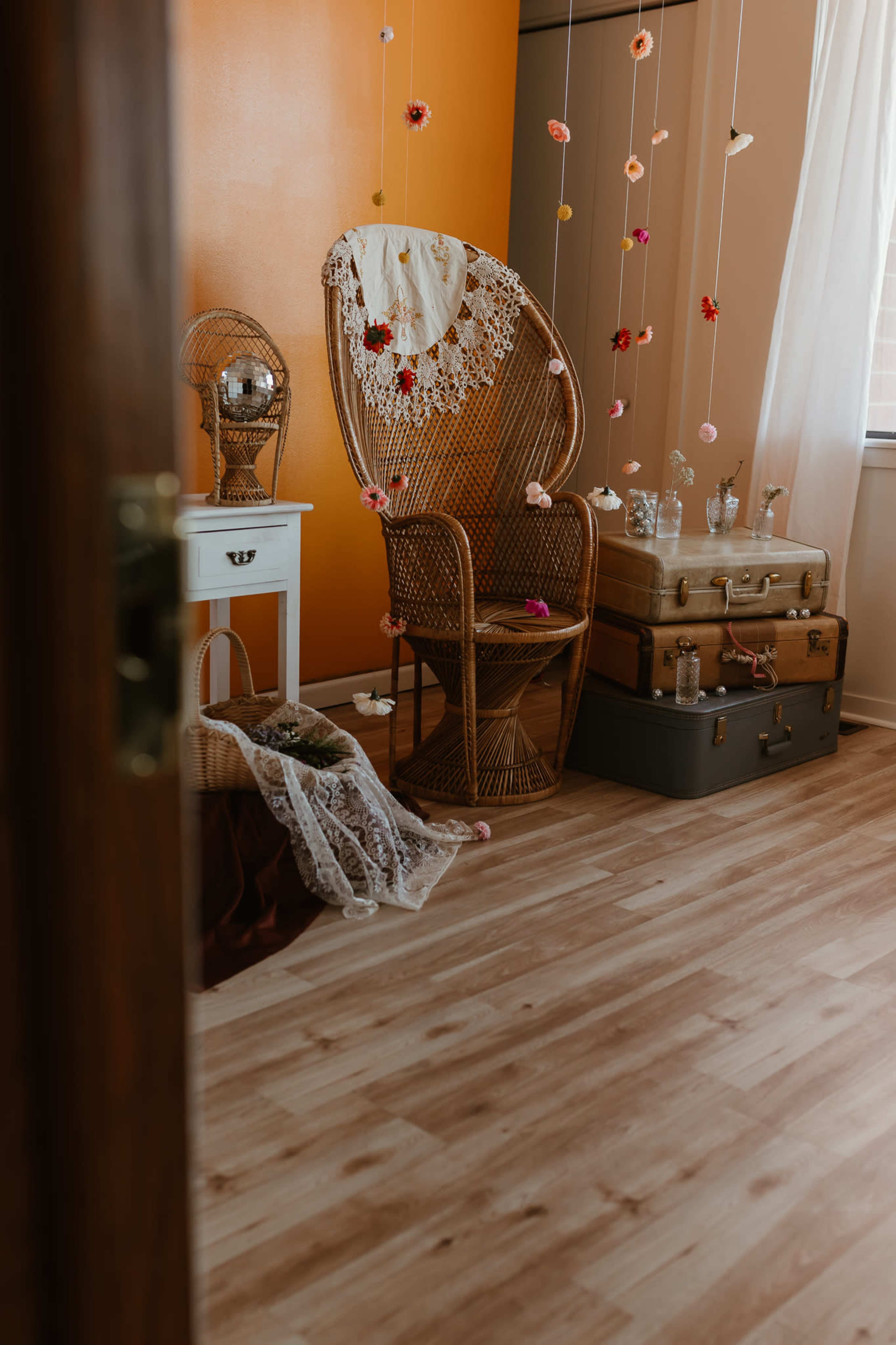 The image shows a cozy room featuring a wicker chair with a lace backrest, a small white nightstand, stacked vintage suitcases, and floral decorations hanging from the ceiling.