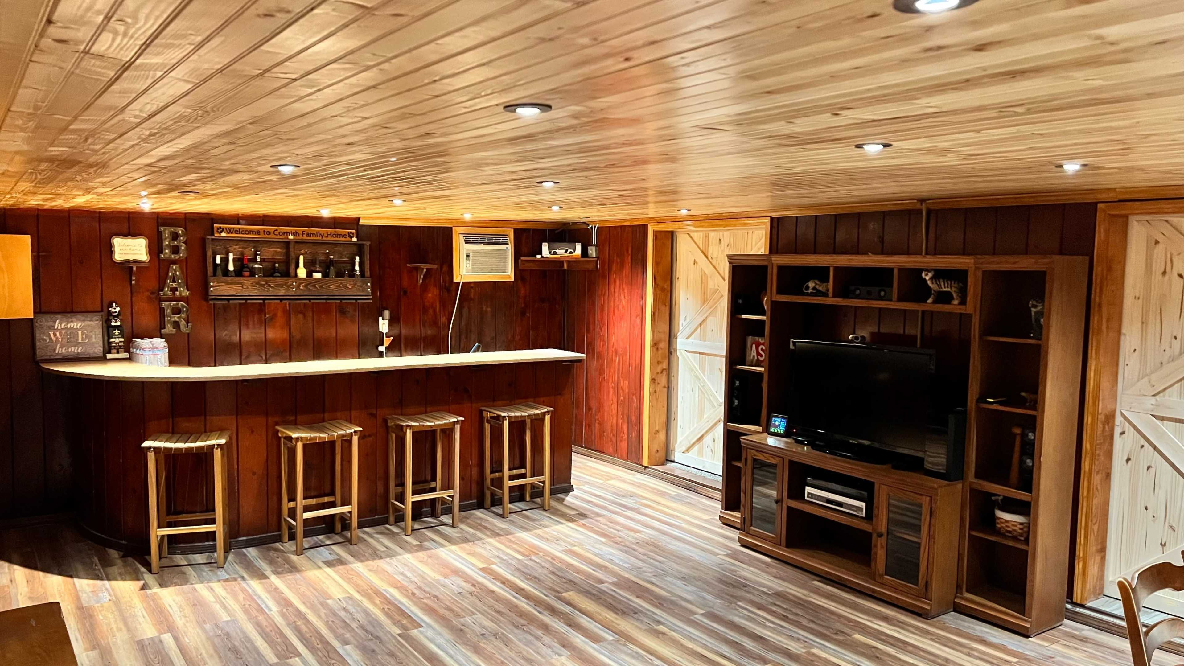A rustic basement bar area featuring wooden paneling, a countertop with bar stools, a television, and shelves for drinks.