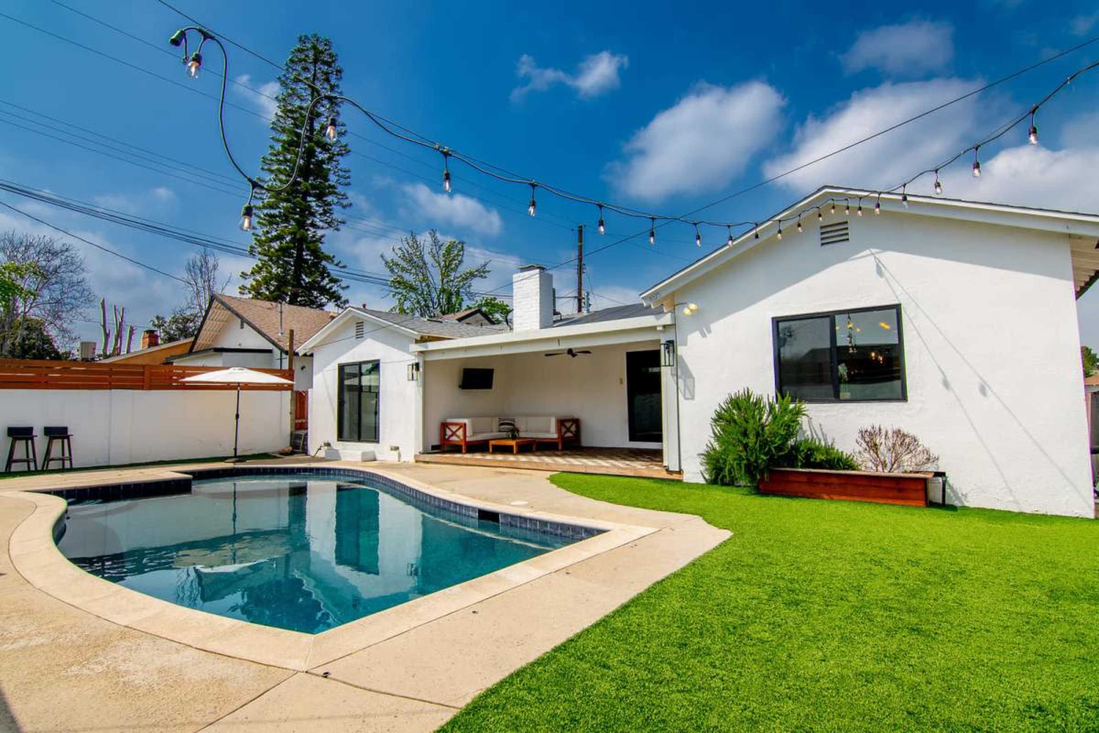 The image shows a backyard with a swimming pool, artificial grass, and a patio area featuring outdoor furniture and string lights above.
