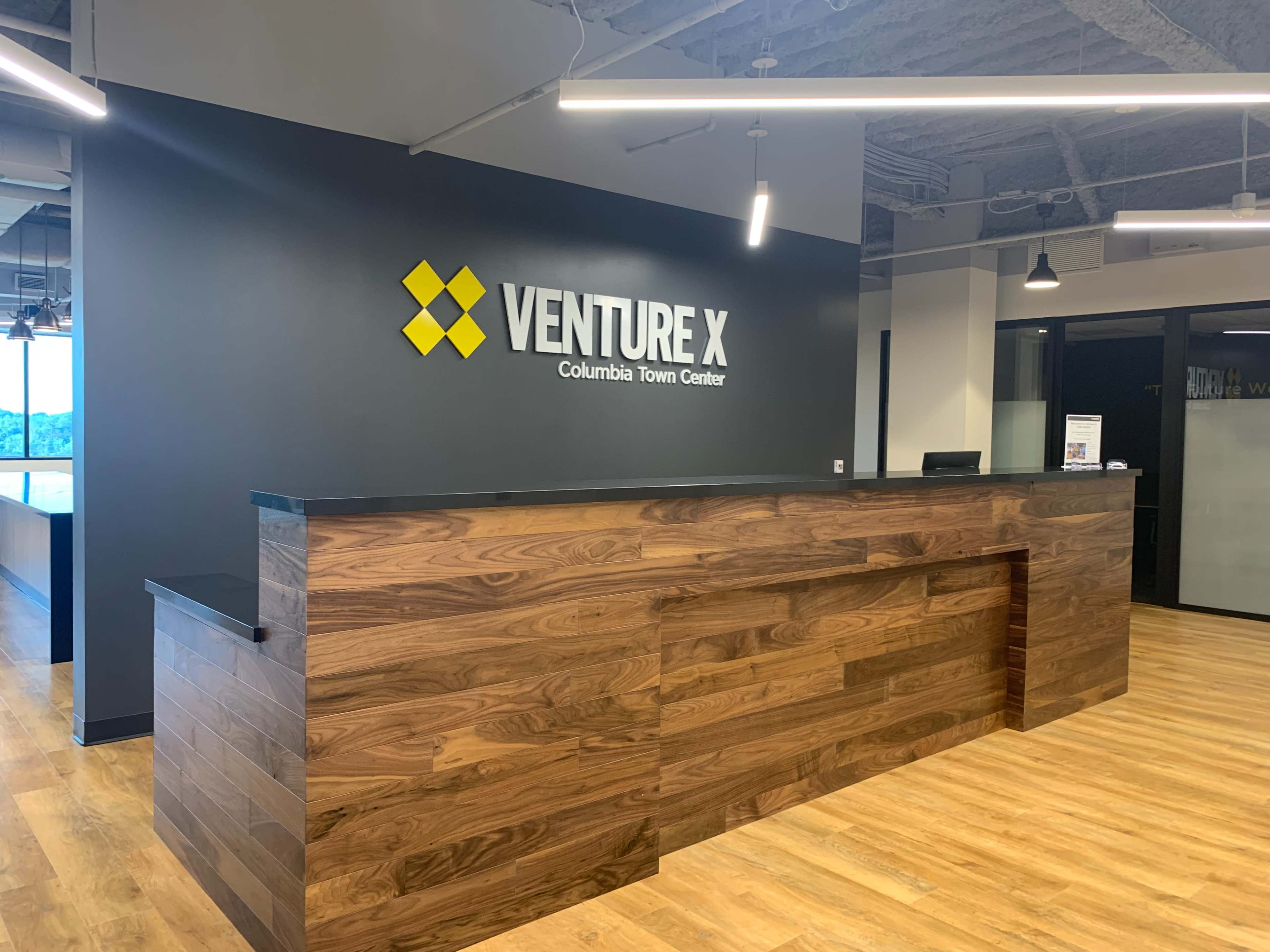 The image shows a reception area with a wooden front desk and a wall featuring the "VENTURE X" logo in yellow and black.