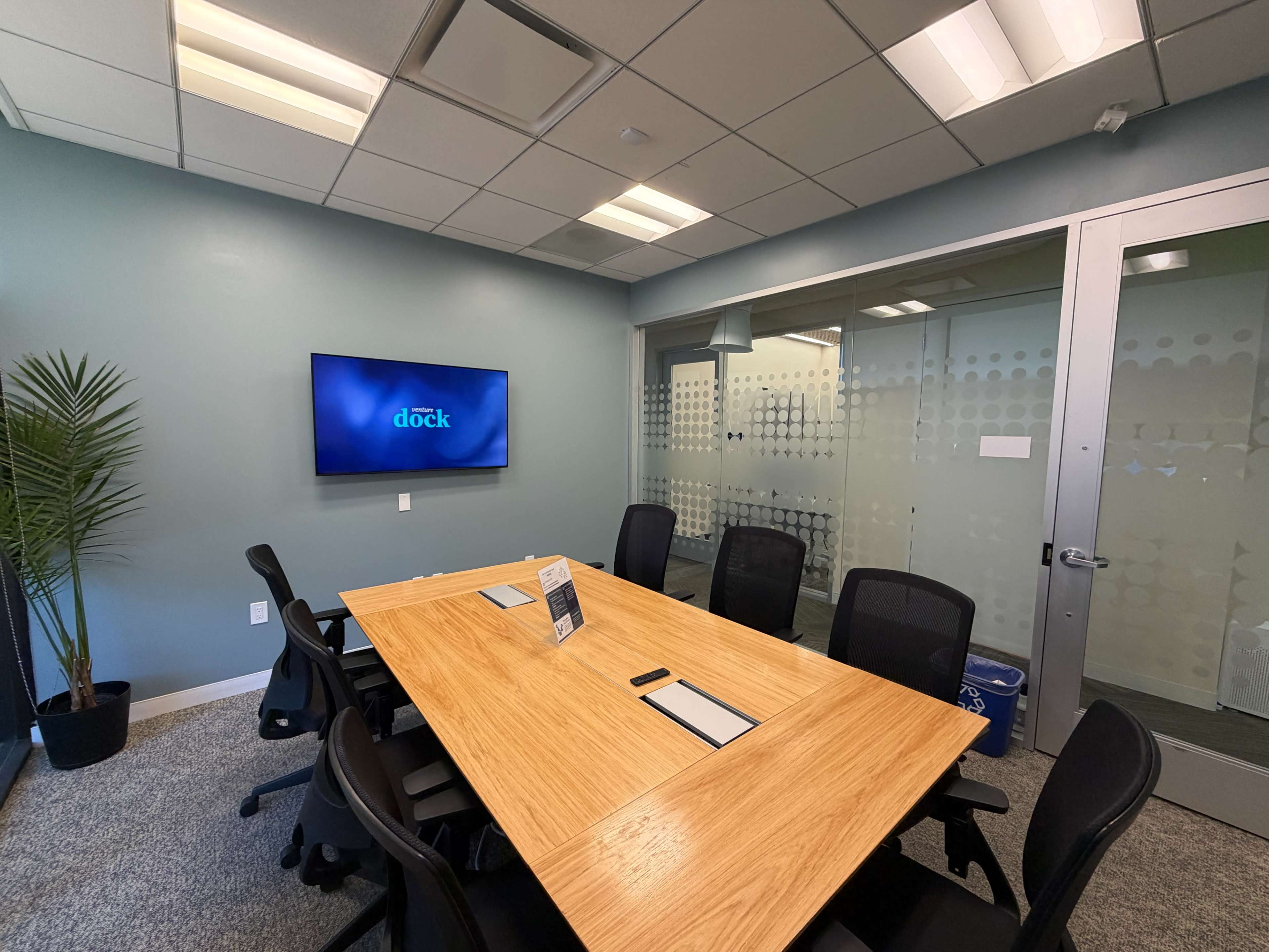 A meeting room features a large wooden table surrounded by black chairs, with a wall-mounted screen displaying the word "dock" and glass panels in the background.