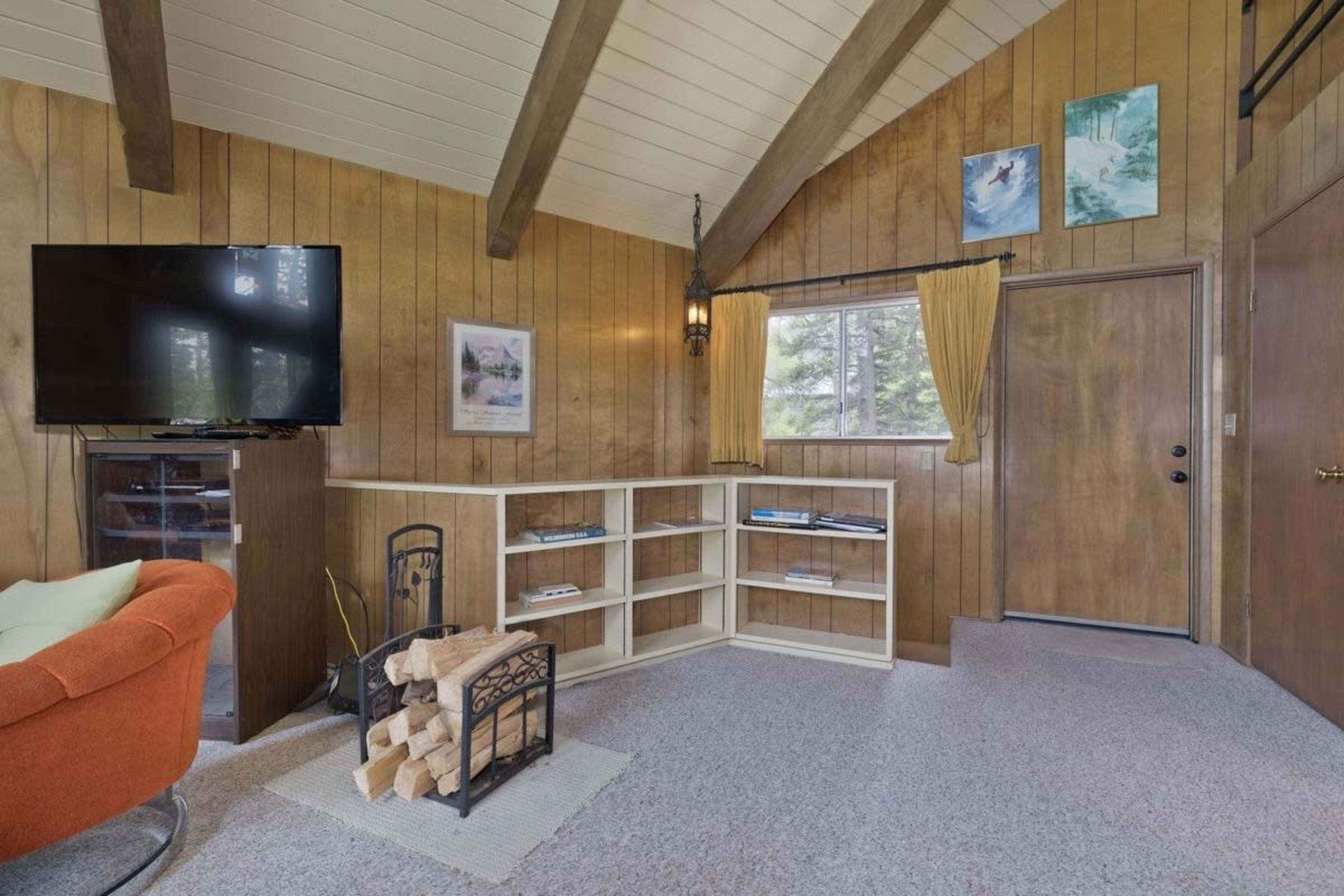 A cozy room features wood-paneled walls, a television mounted on one side, a shelving unit, a door, and a stack of firewood on the floor.
