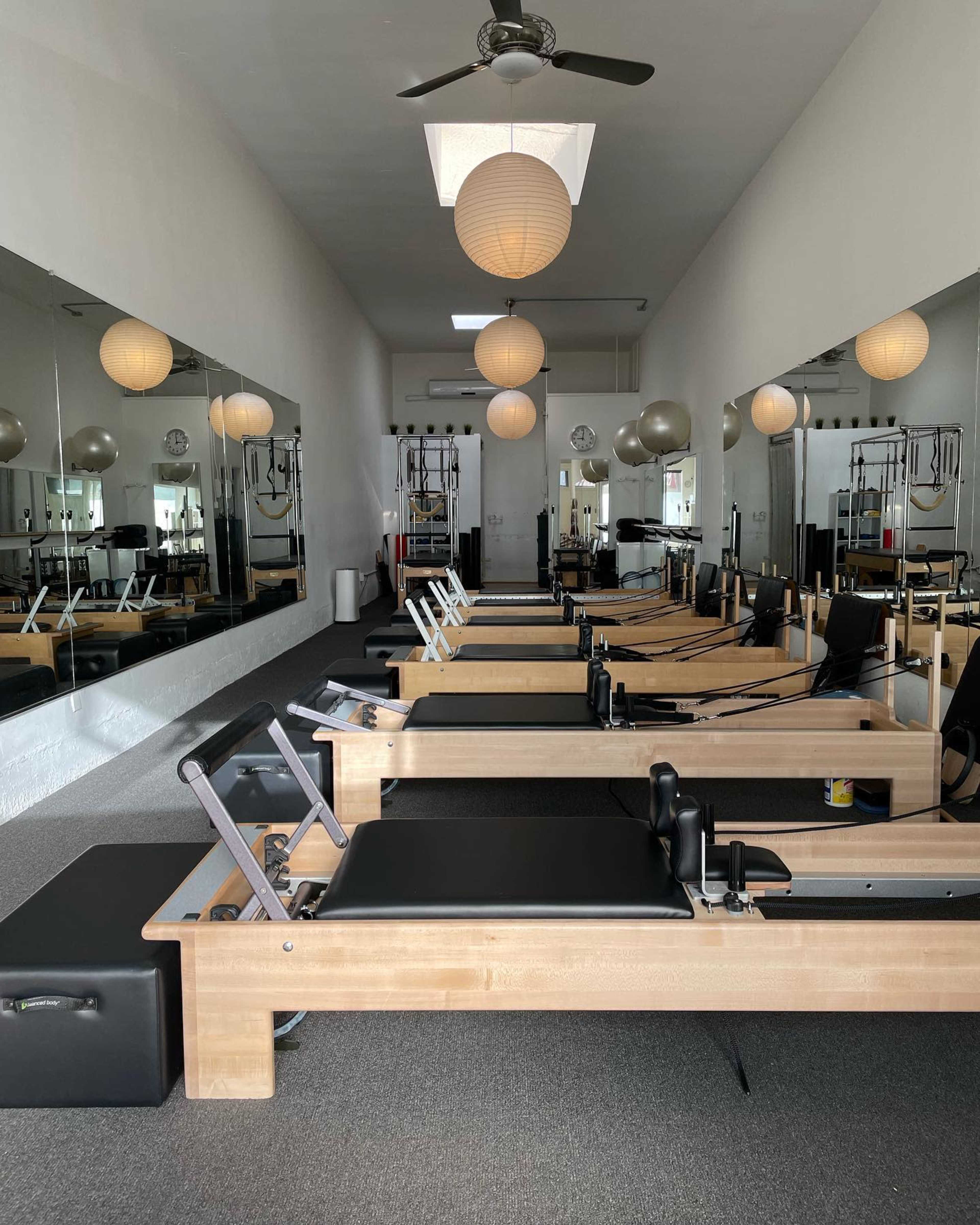A well-lit Pilates studio featuring multiple reformer machines aligned in rows, with large mirrors reflecting the space and pendant lights hanging from the ceiling.
