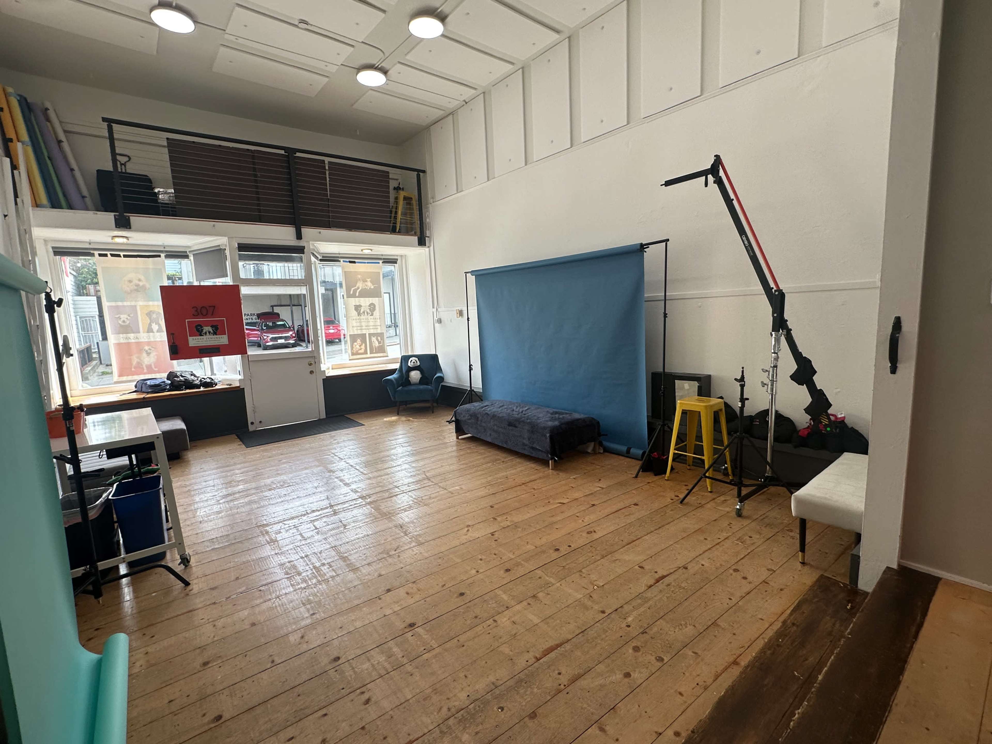 The image shows a spacious studio with wooden floors, a blue backdrop, a single chair, and various photography equipment, illuminated by natural light coming through large windows.