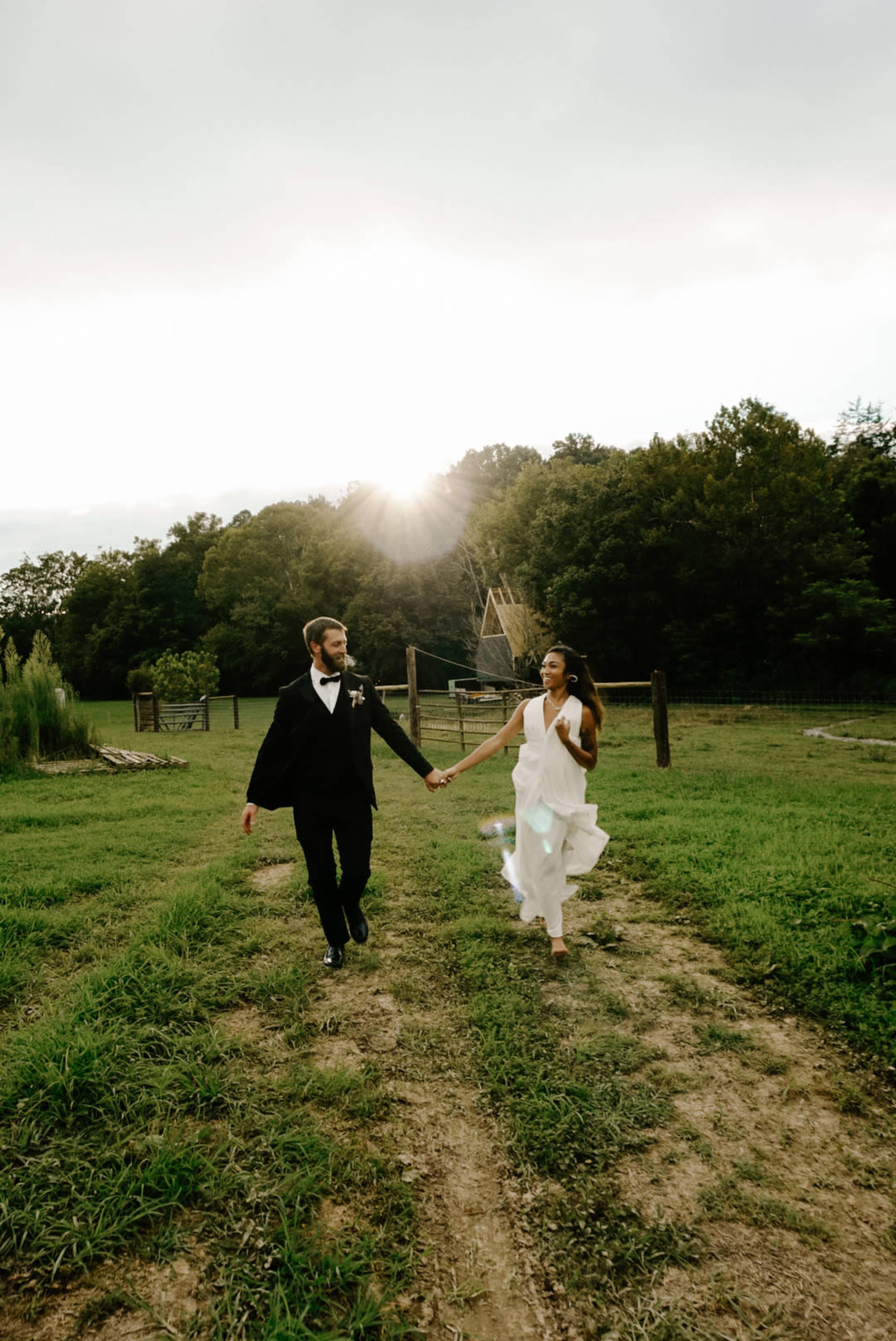 A man in a black suit and a woman in a white dress hold hands while walking along a dirt path in a grassy field, with trees and a sunbeam in the background.