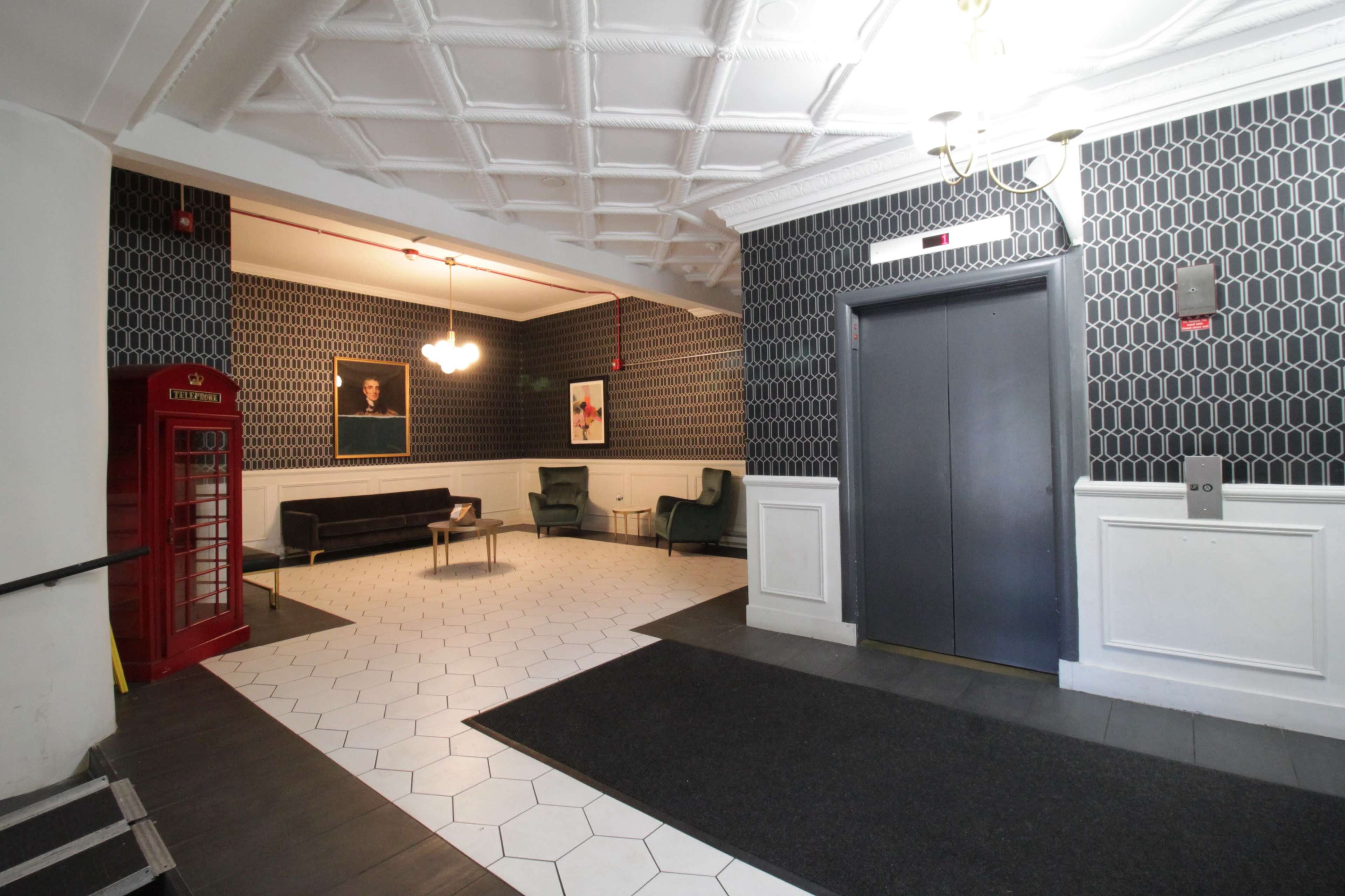 The image shows a lobby area featuring an elevator, a red phone booth, a sofa, two chairs, and patterned wallpaper.