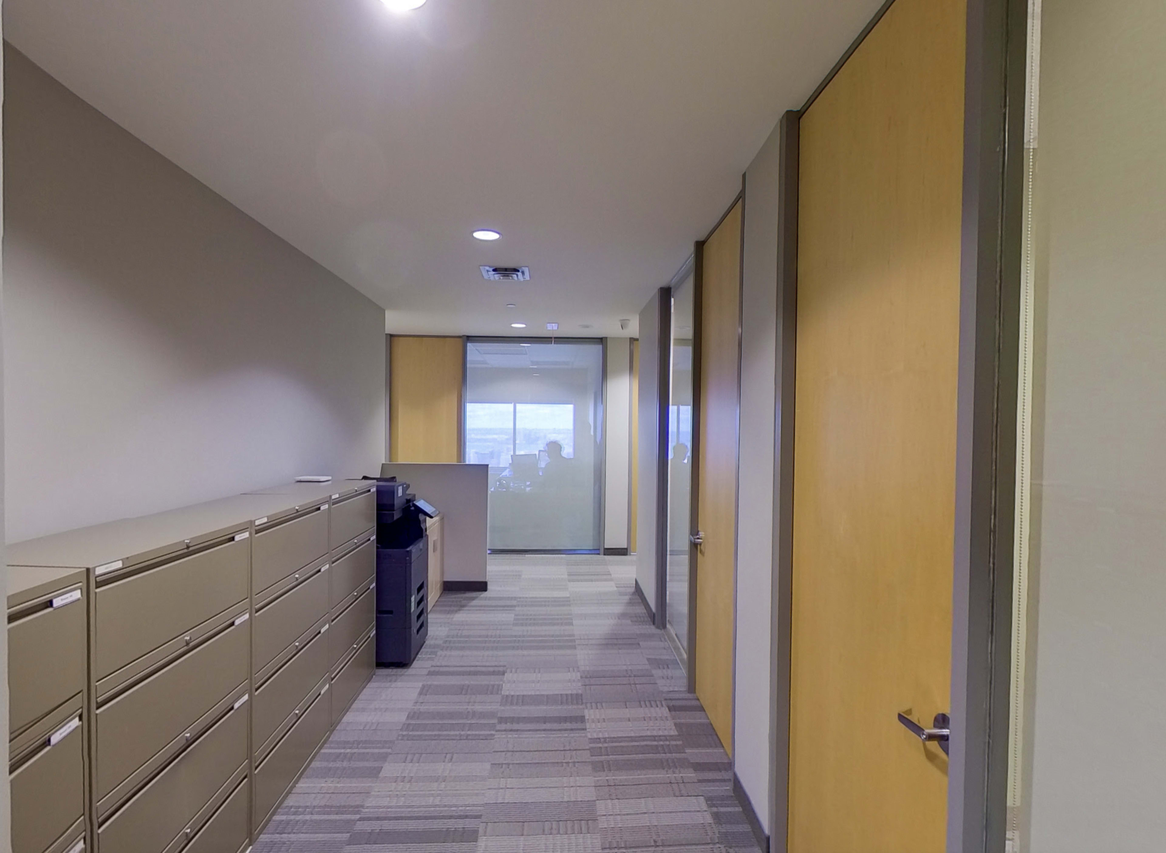 The image shows a contemporary office hallway with gray cabinets on one side and glass-paneled meeting rooms on the other.