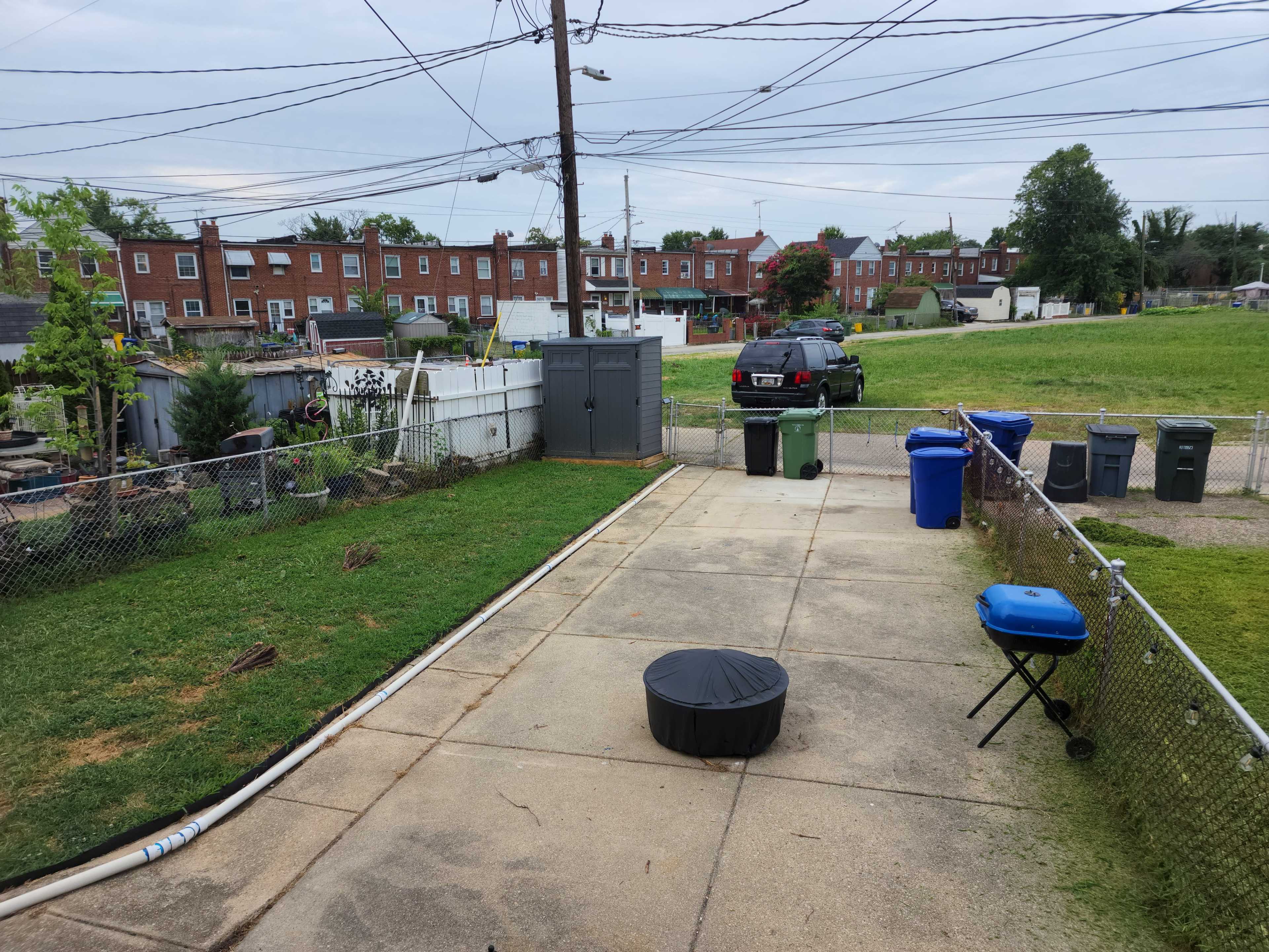 The image shows a concrete backyard area with a circular black cover, a blue grill, trash bins in various colors, and a fenced yard with residential buildings and grass in the background.