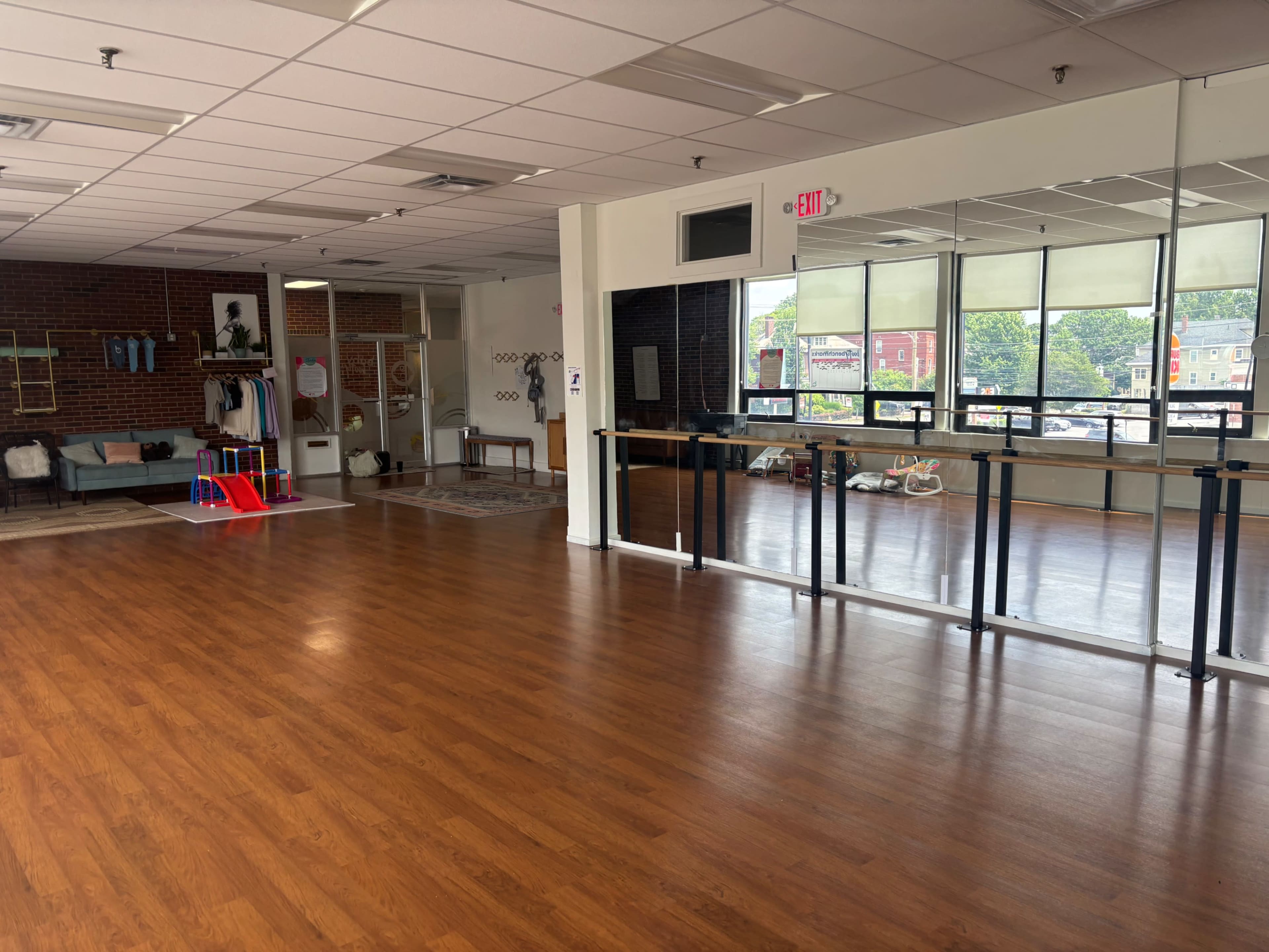 The image shows an empty studio space with wooden flooring, large mirrors on one wall, and a play area with colorful equipment in the corner.