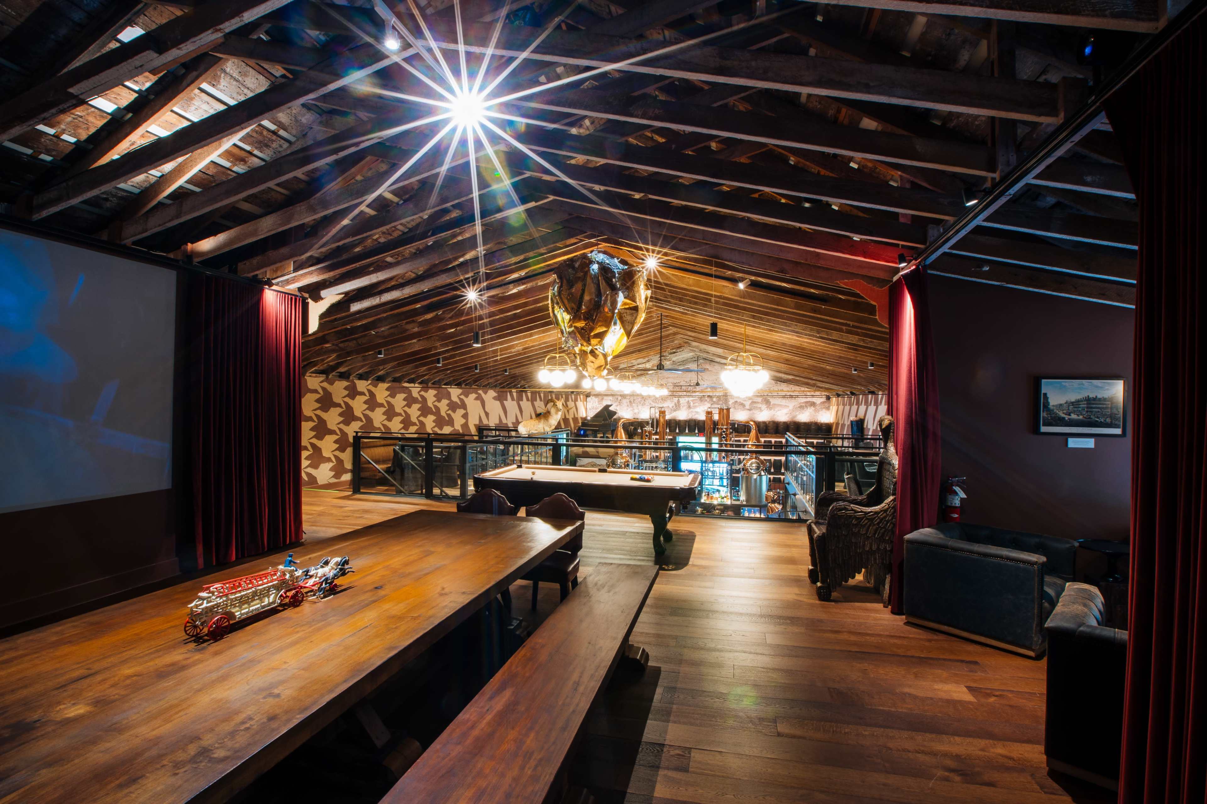 The image shows a spacious interior room with wooden beams, a large dining table, and a multi-level area featuring a bar and seating.