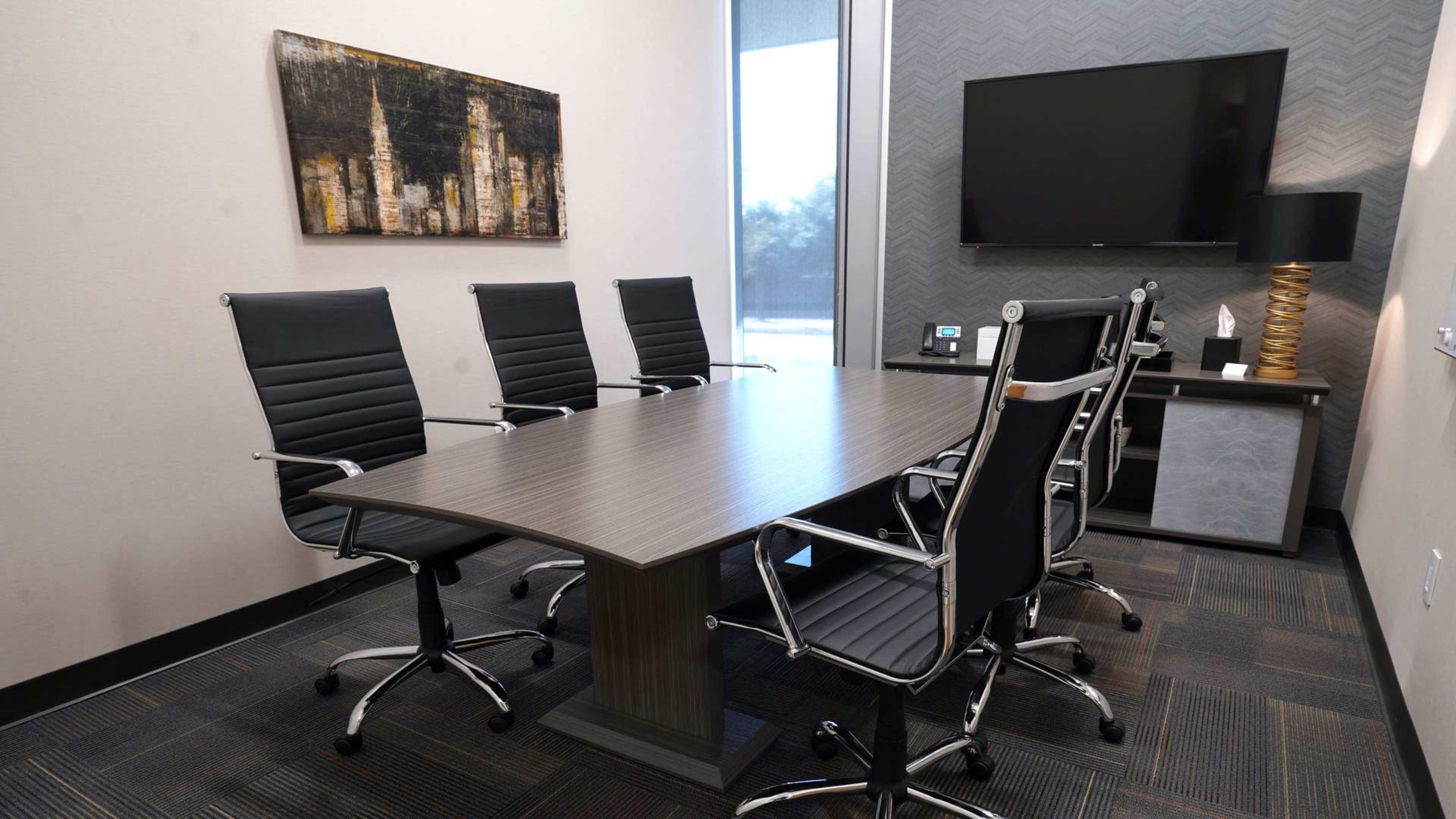 A modern conference room features a large rectangular table surrounded by six black ergonomic chairs, with a television mounted on the wall and a decorative painting in the background.