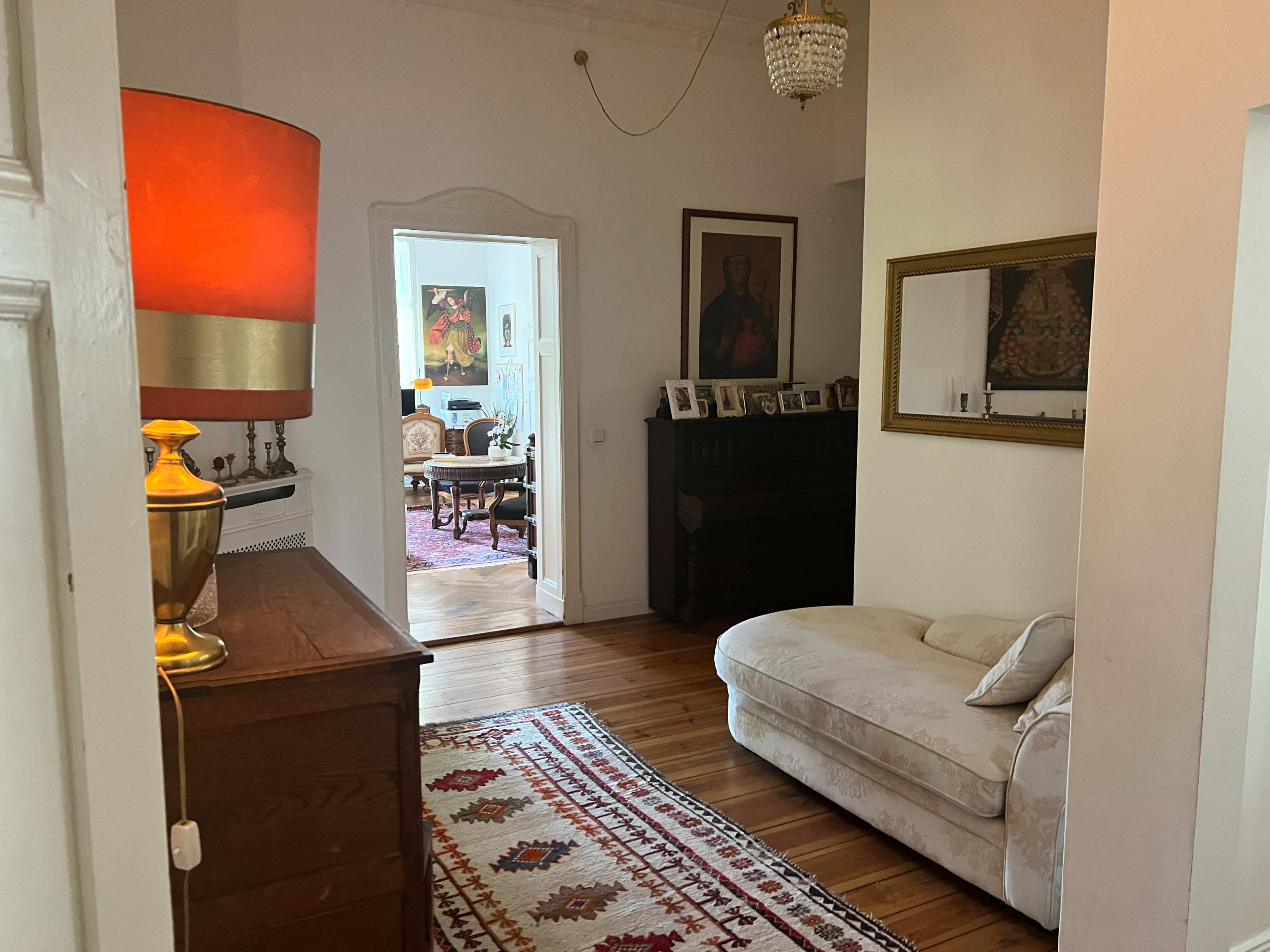 A cozy interior hallway leading to a bright living area, with a sofa, a decorative rug, and framed photos on a wooden chest.