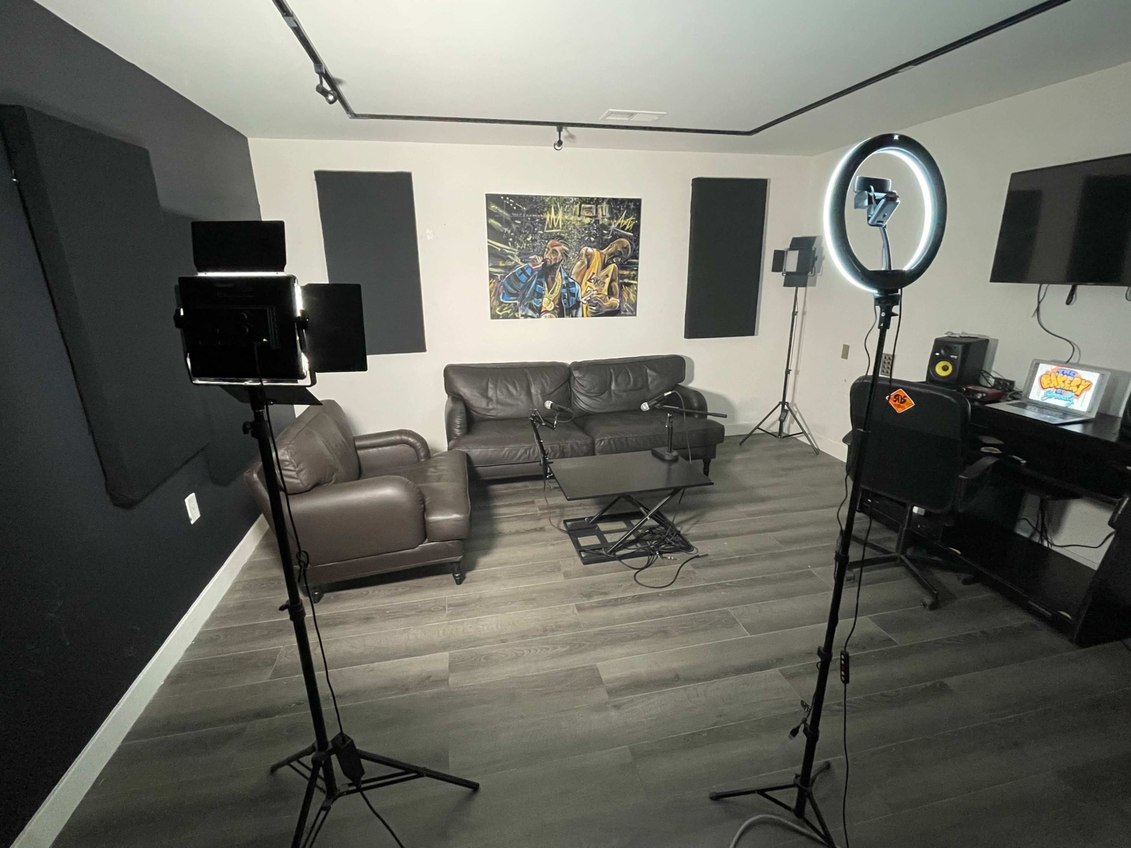 A modern, minimalist studio setup with a couch, a coffee table, and lighting equipment arranged around a wall featuring a large art piece.