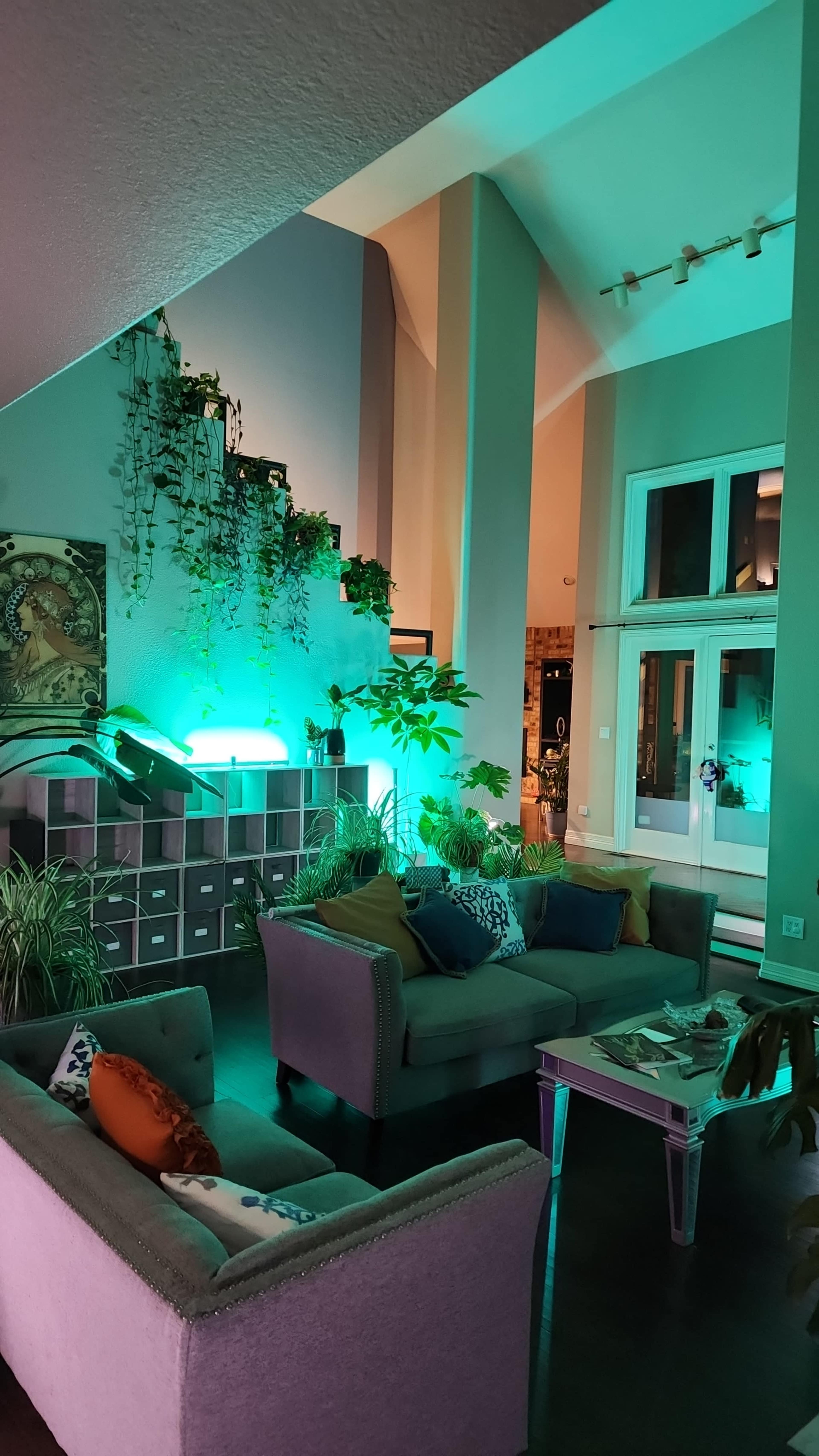 The image shows a well-lit living room with green ambient lighting, plants, and a staircase in the background.