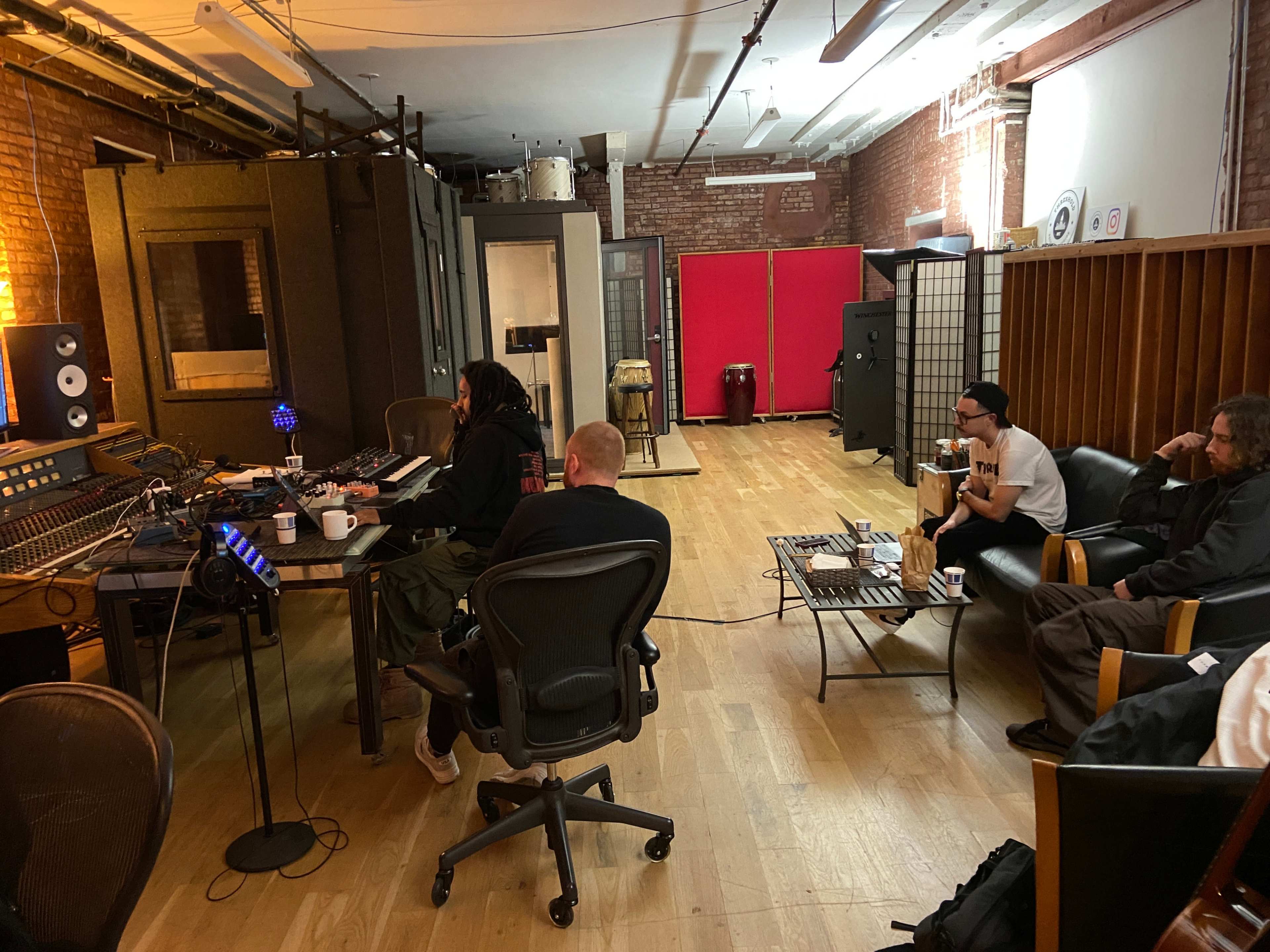 The image shows a recording studio with three individuals: two seated at a sound mixing console and one sitting on a couch in a lounge area, surrounded by musical equipment.