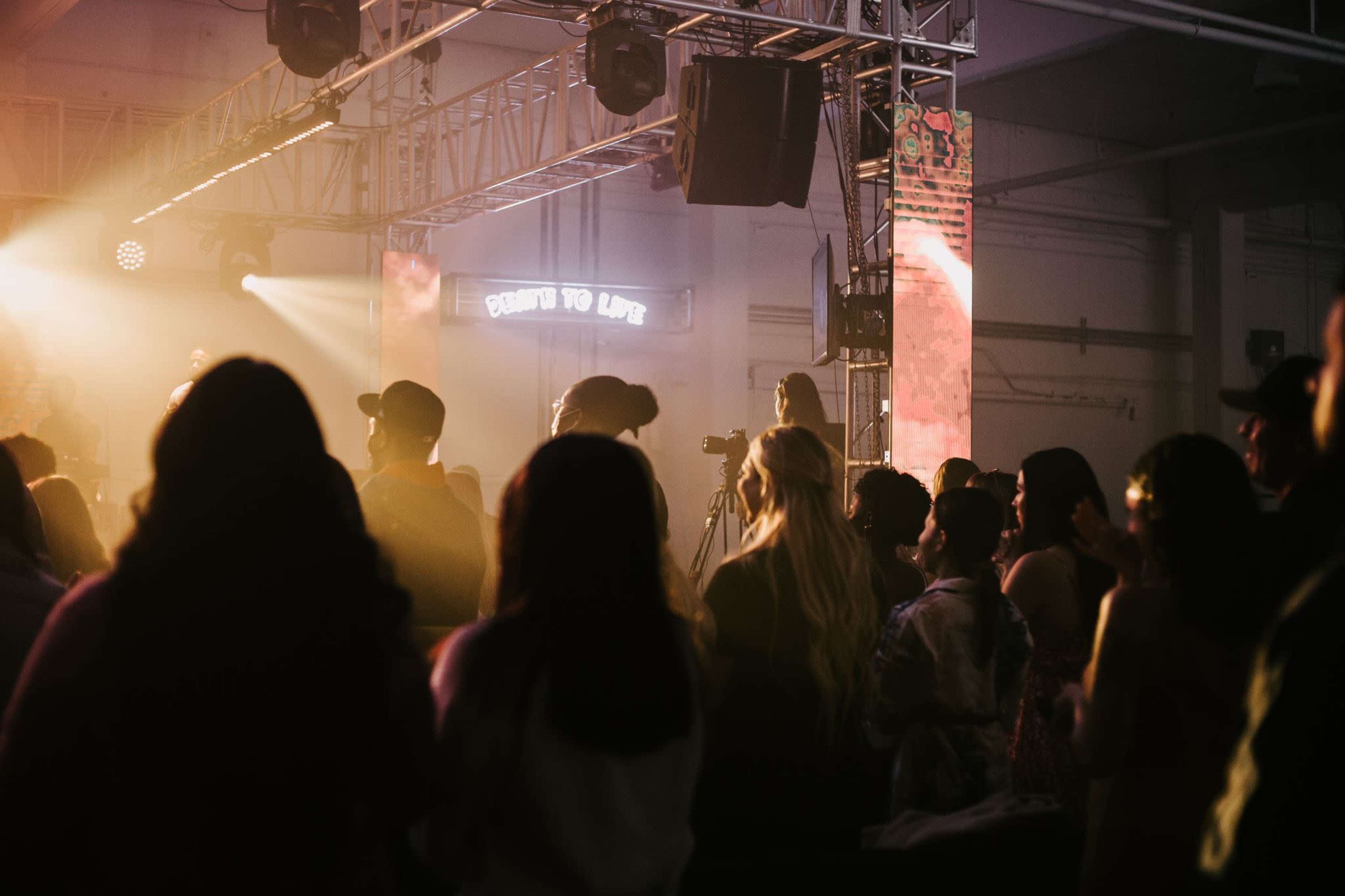 A crowd of people stands in a dimly lit venue while a performance takes place on stage.